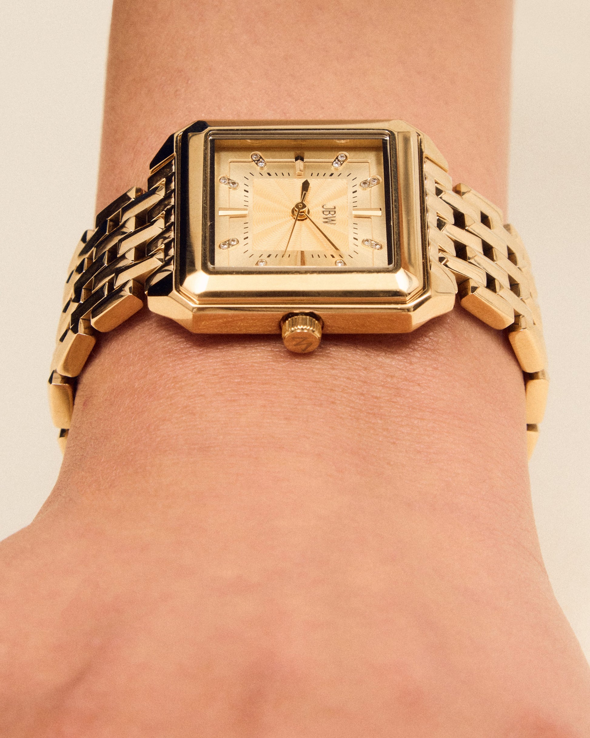 A close-up of the JBW Watches Astra | J6410B, a gold square silhouette watch with a guilloché-textured dial and linked band, on a wrist displaying the time at around 7:05.