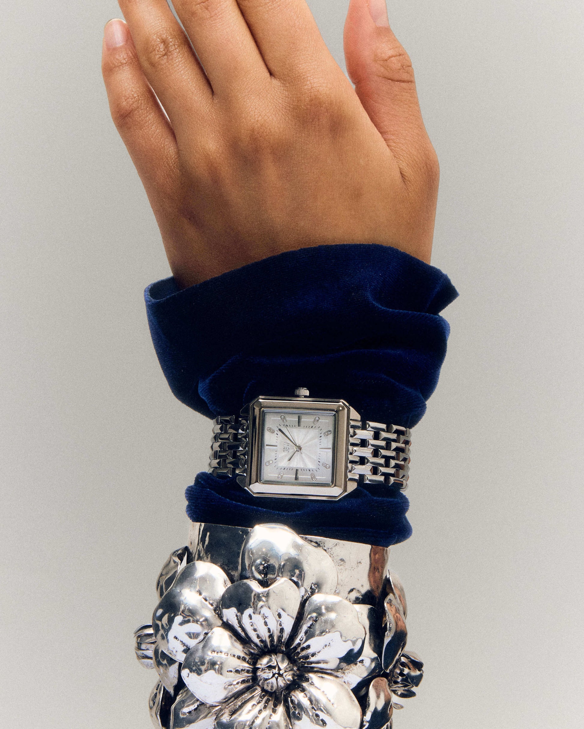 A hand wearing the JBW Watches Astra | J6410A, a square silver wristwatch, styled with a chunky silver floral bracelet and a deep blue velvet sleeve, is displayed against a plain background.
