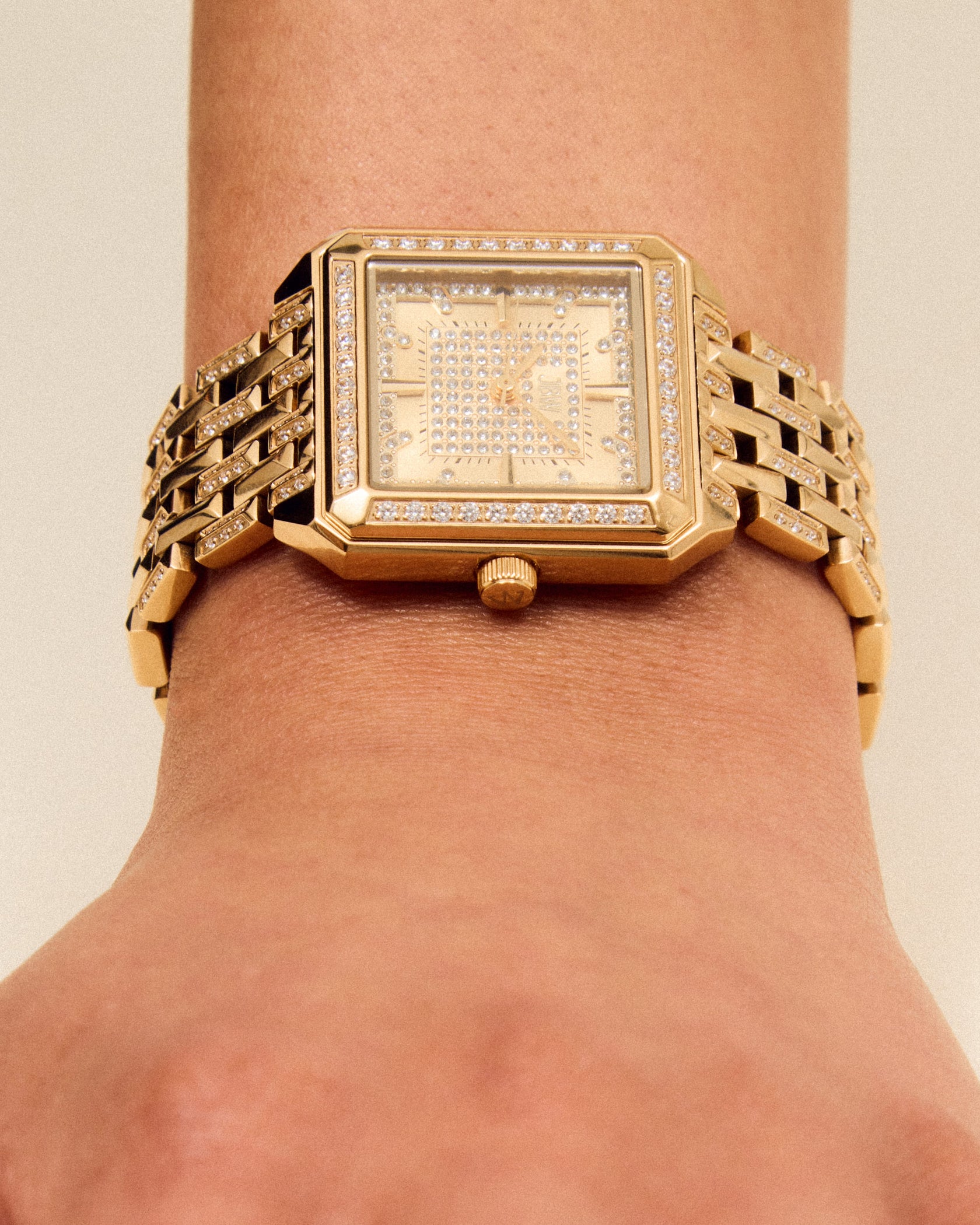 A close-up of a wrist wearing the JBW Watches Astra | J6411B, a gold square watch with a linked band and jeweled bezel. The pavé-grid dial enhances elegance as the relaxed hand rests on a neutral background.