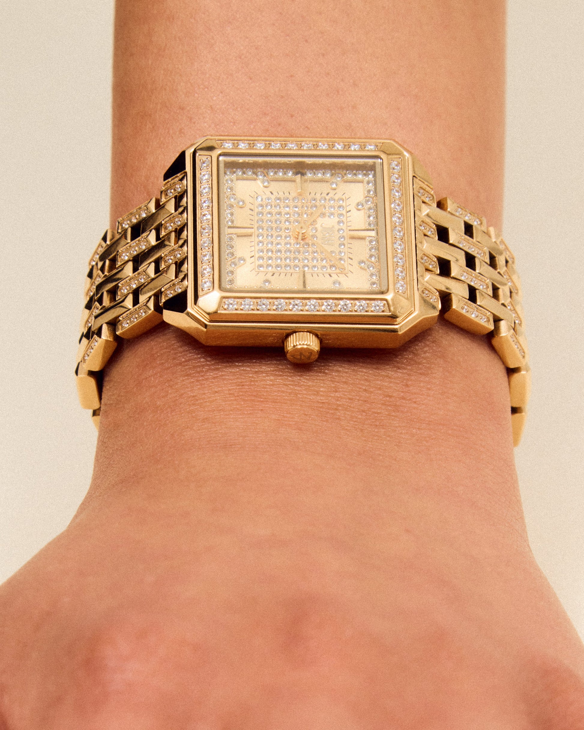 A close-up of a wrist wearing the JBW Watches Astra | J6411B, a gold square watch with a linked band and jeweled bezel. The pavé-grid dial enhances elegance as the relaxed hand rests on a neutral background.