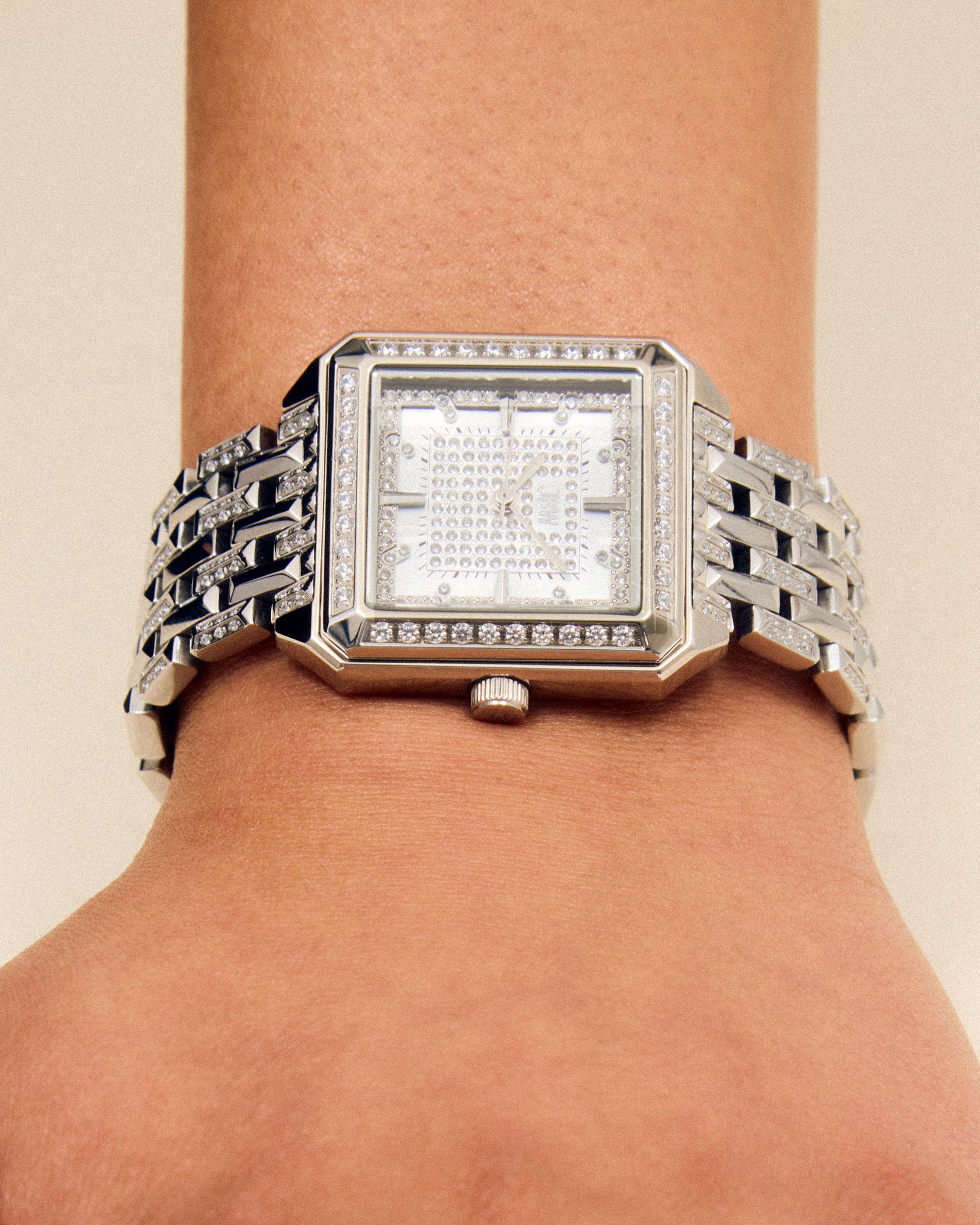 A close-up of a wrist wearing the JBW Watches Astra | J6411A, a silver square watch with a jeweled face and bracelet-style band, set against a light beige background.