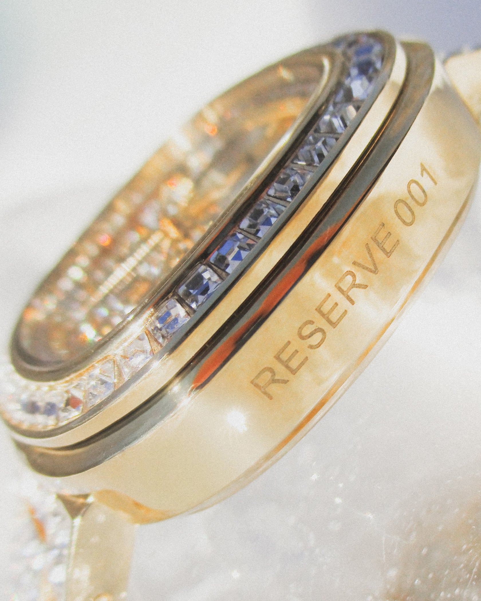 Close-up of the JBW Watches RESERVE Coast Chandelier | R6397D, featuring RESERVE 001 engraved on its gold side and a blue gemstone bezel, paired with a gold-tone bracelet. The watch face appears slightly blurred in the background.