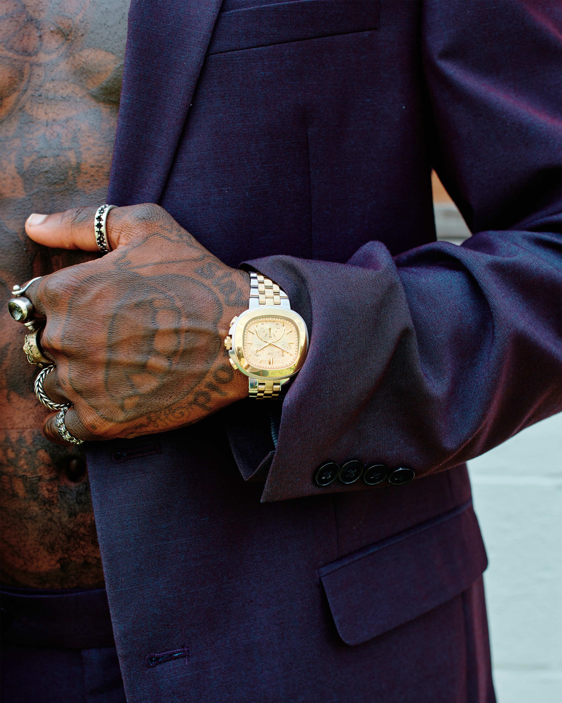 A person in a dark suit displays a tattooed chest and hand adorned with rings, wearing the JBW Watches Coast | J6397C gold chronograph watch. Their hand rests on their chest, highlighting lab-grown diamond-set jewelry and the luxury timepiece.