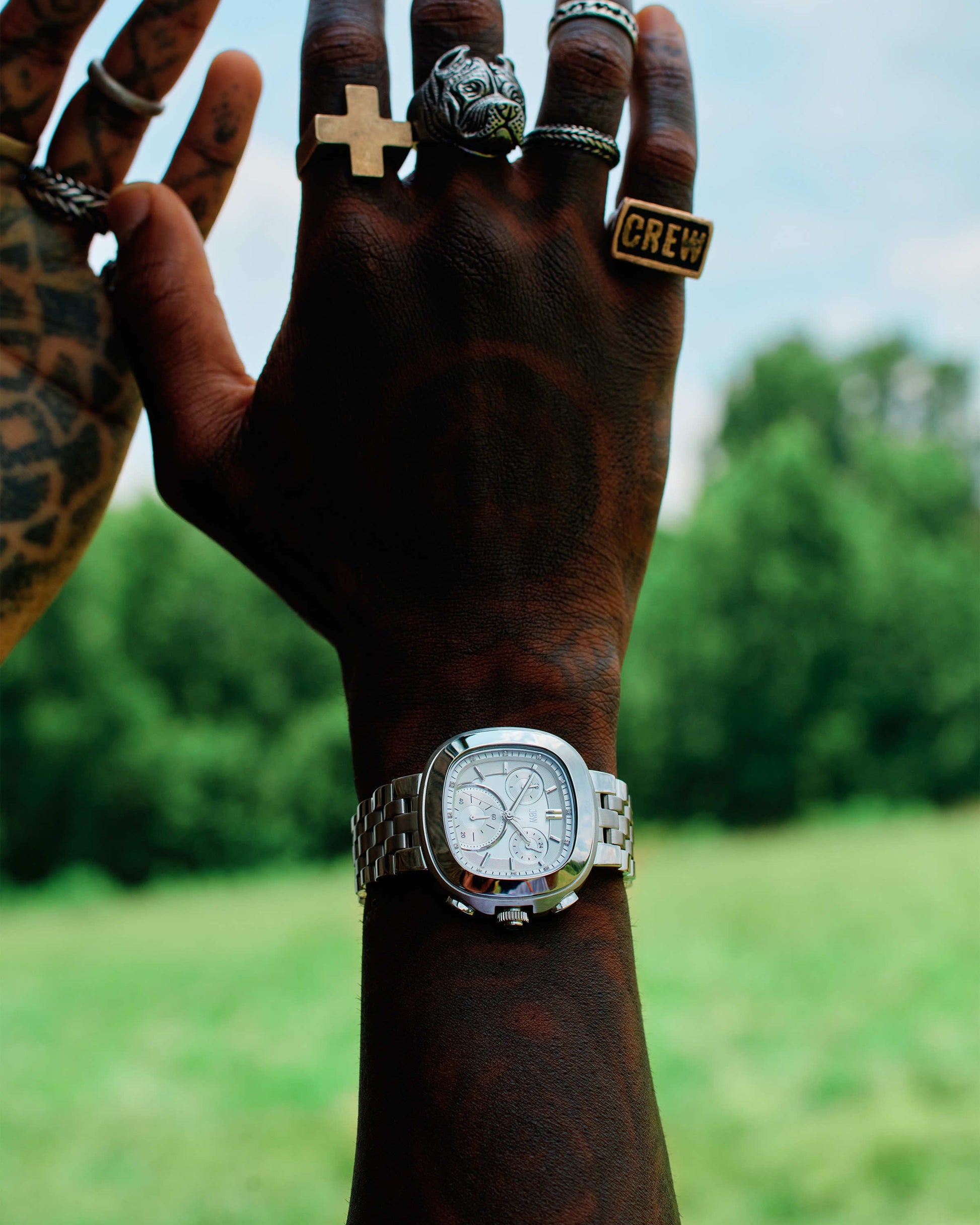 A tattooed hand raised outdoors, adorned with rings (cross, lion, CREW), wears the 38mm JBW Watches Coast | J6397B stainless steel watch against a blurred leafy green background.