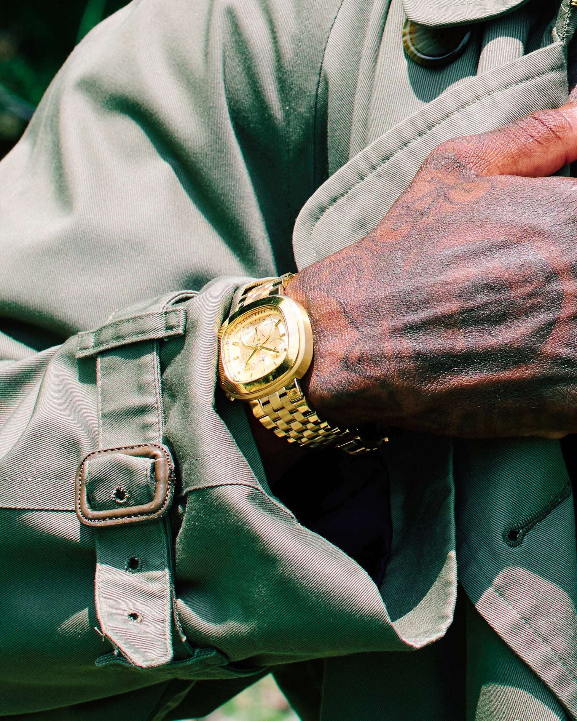 A close-up of a tattooed hand wearing the JBW Watches Coast | J6397A gold and stainless steel watch with an olive green trench coat, holding the lapel. The details of both the watch and coat are clearly visible.