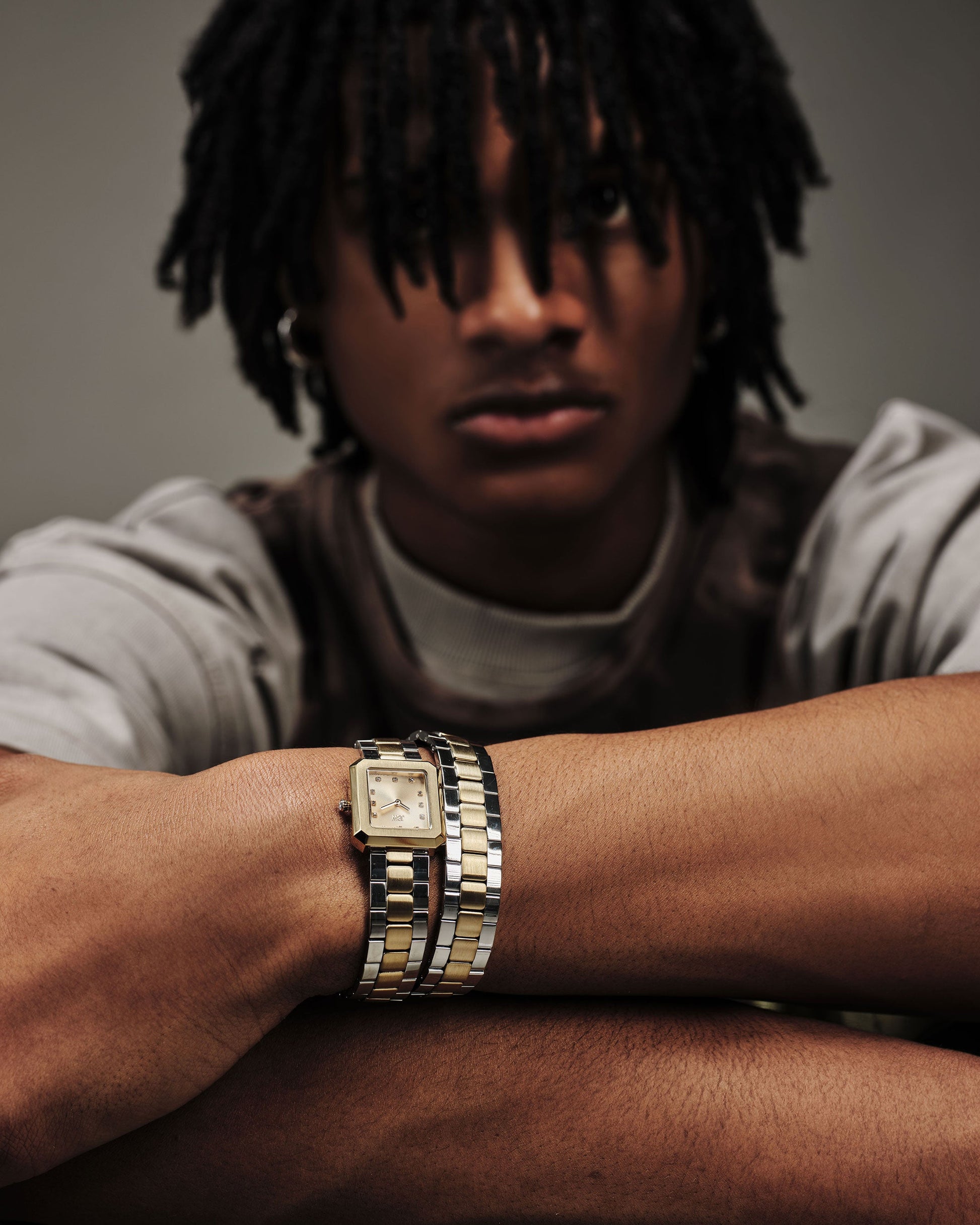 A person with medium-length dreadlocks, arms crossed, gazes forward while wearing the JBW Watches Arc Double (Sample Sale) | J6390K-GA. The focus is on the watch; their face appears partially blurred in the background.