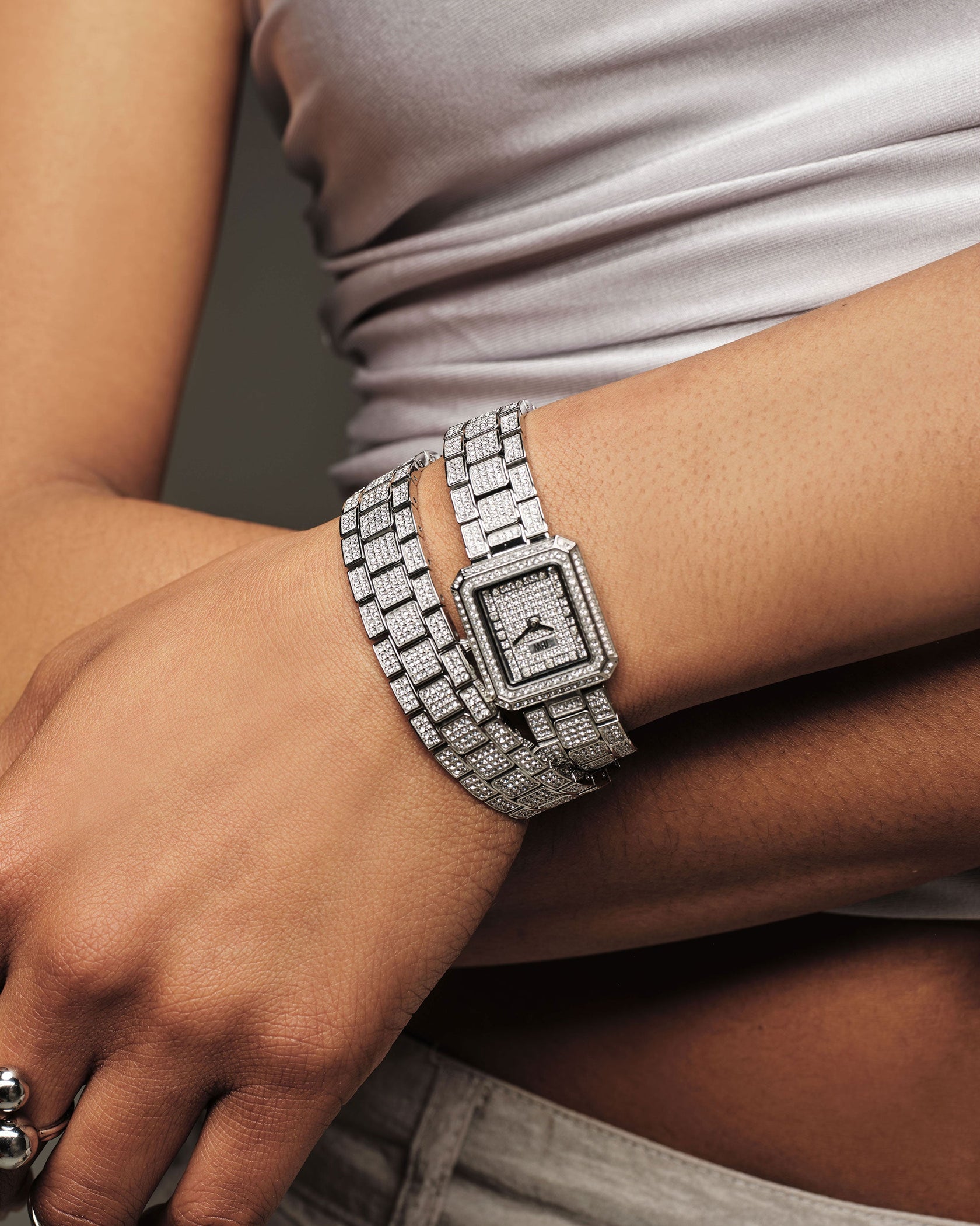A person wears the JBW Watches Arc Double | J6390D, featuring a crystal-studded bezel, with two diamond-studded bracelets, paired with a light gray satin top and light-colored pants.