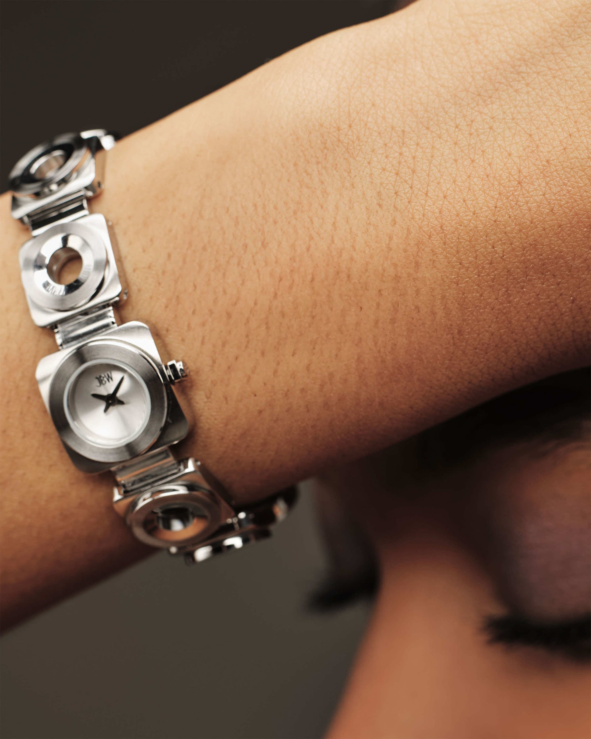 A close-up shows a person wearing the JBW Watches Chasm (Sample Sale) | J6398B-SC, a modern stainless steel watch with a geometric square design, highlighted against a softly blurred background.