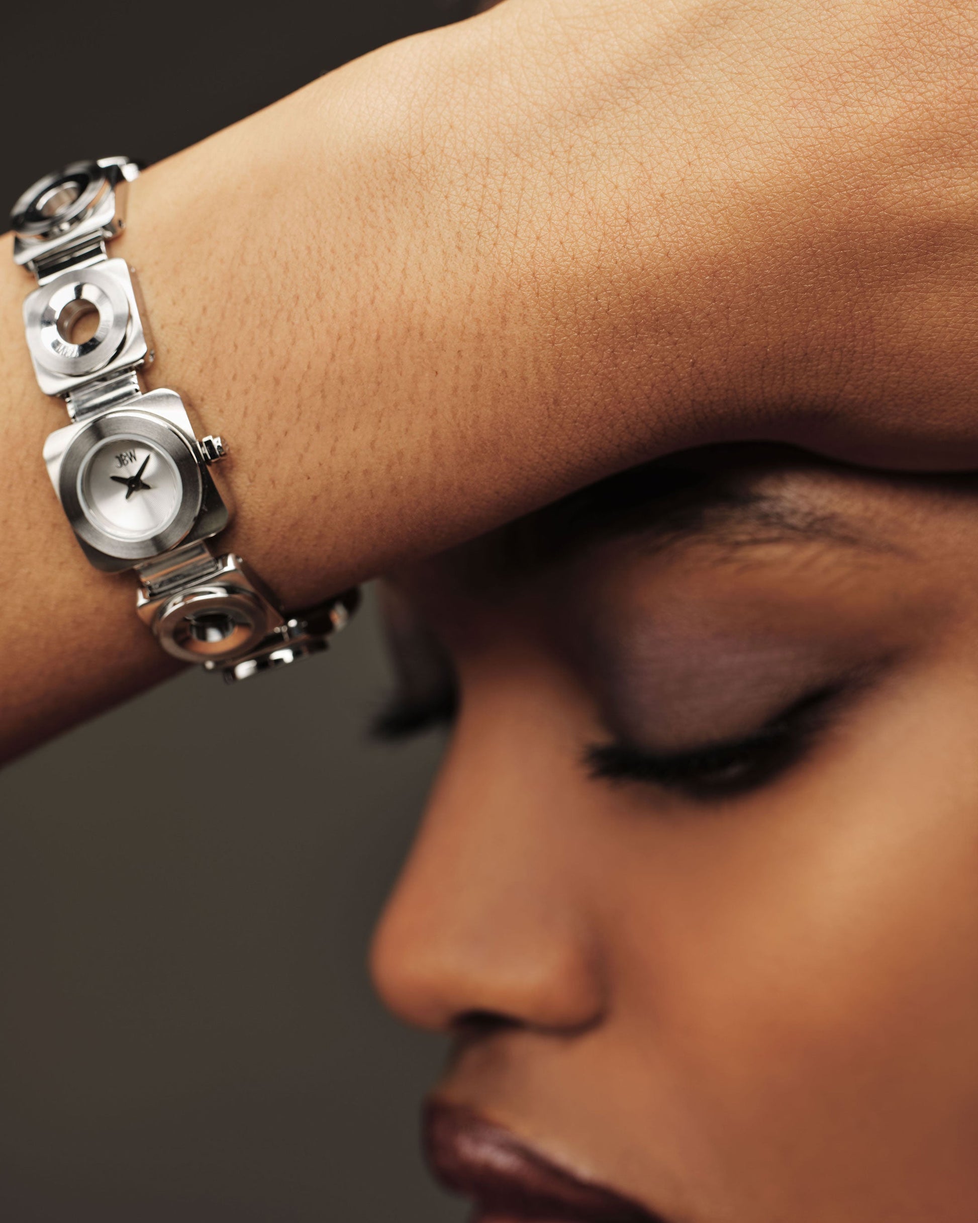 With closed eyes, a person rests their forehead on their arm, highlighting the JBW Watches Chasm Petite (Sample Sale) | J6400B-SC—a silver timepiece with a geometric design and stainless steel bracelet—captured in soft light.