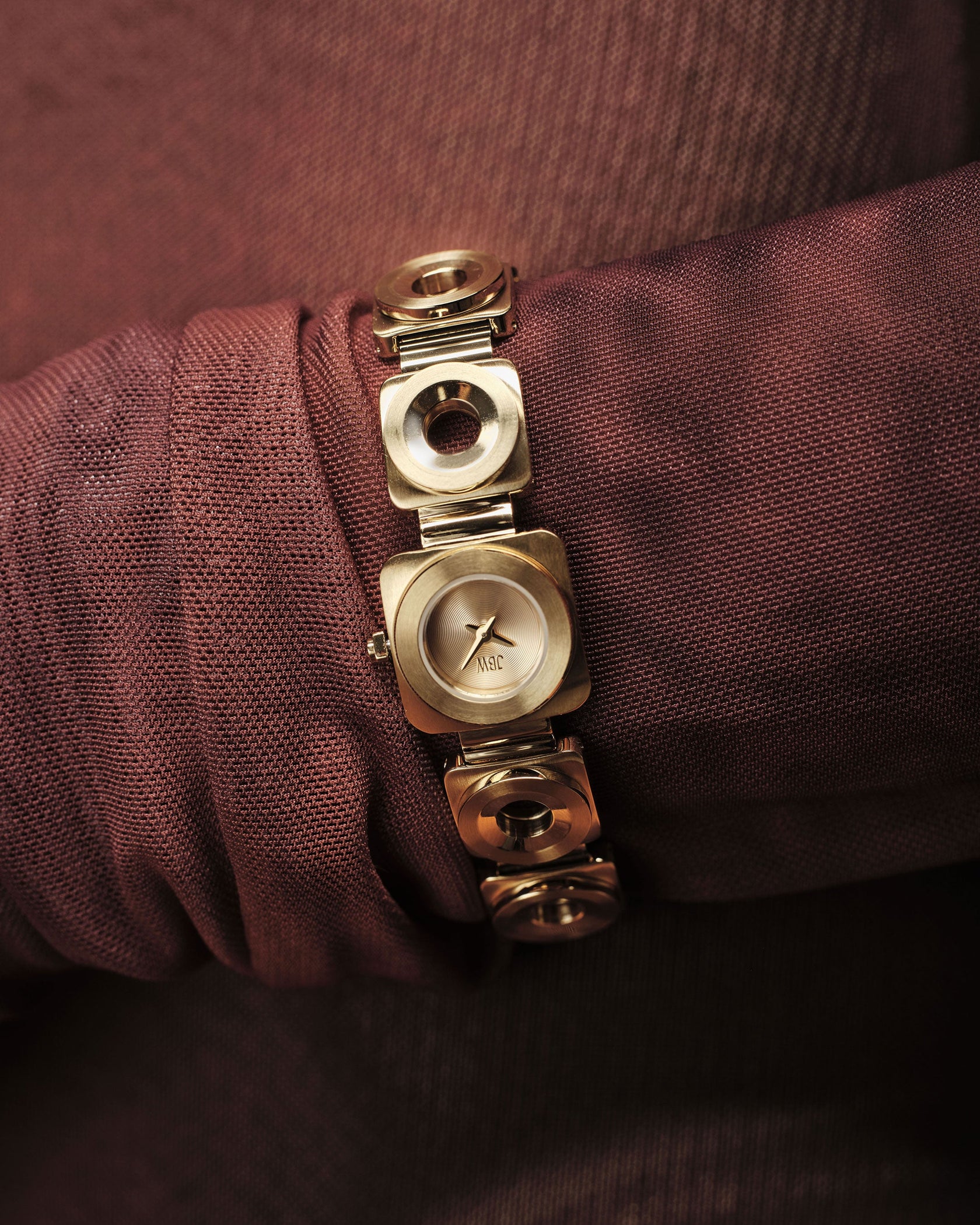 A close-up of the JBW Watches Chasm Petite | J6400A, an 18k Gold-plated watch with a square face and circular links, elegantly worn over sheer, textured, reddish-brown fabric.