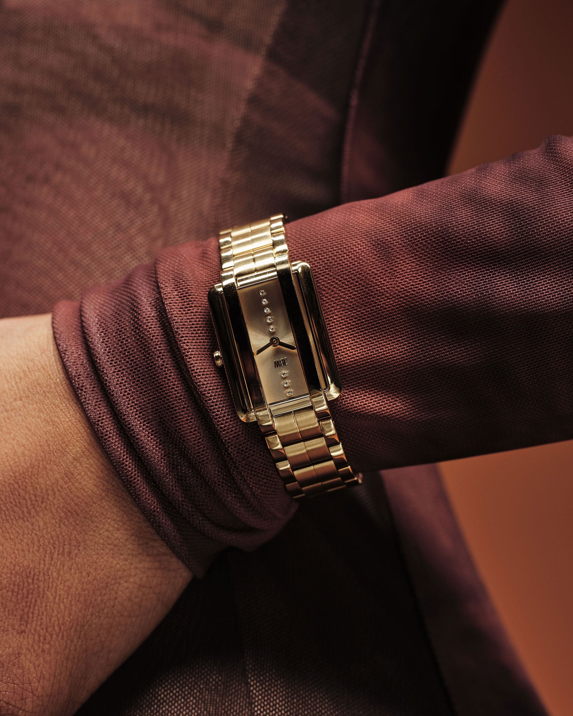 A close-up of a person wearing the JBW Watches Mink Petite | J6395A, a gold luxury watch with a rectangular black face, paired with a sheer maroon long-sleeve top. The softly blurred background is filled with warm tones.