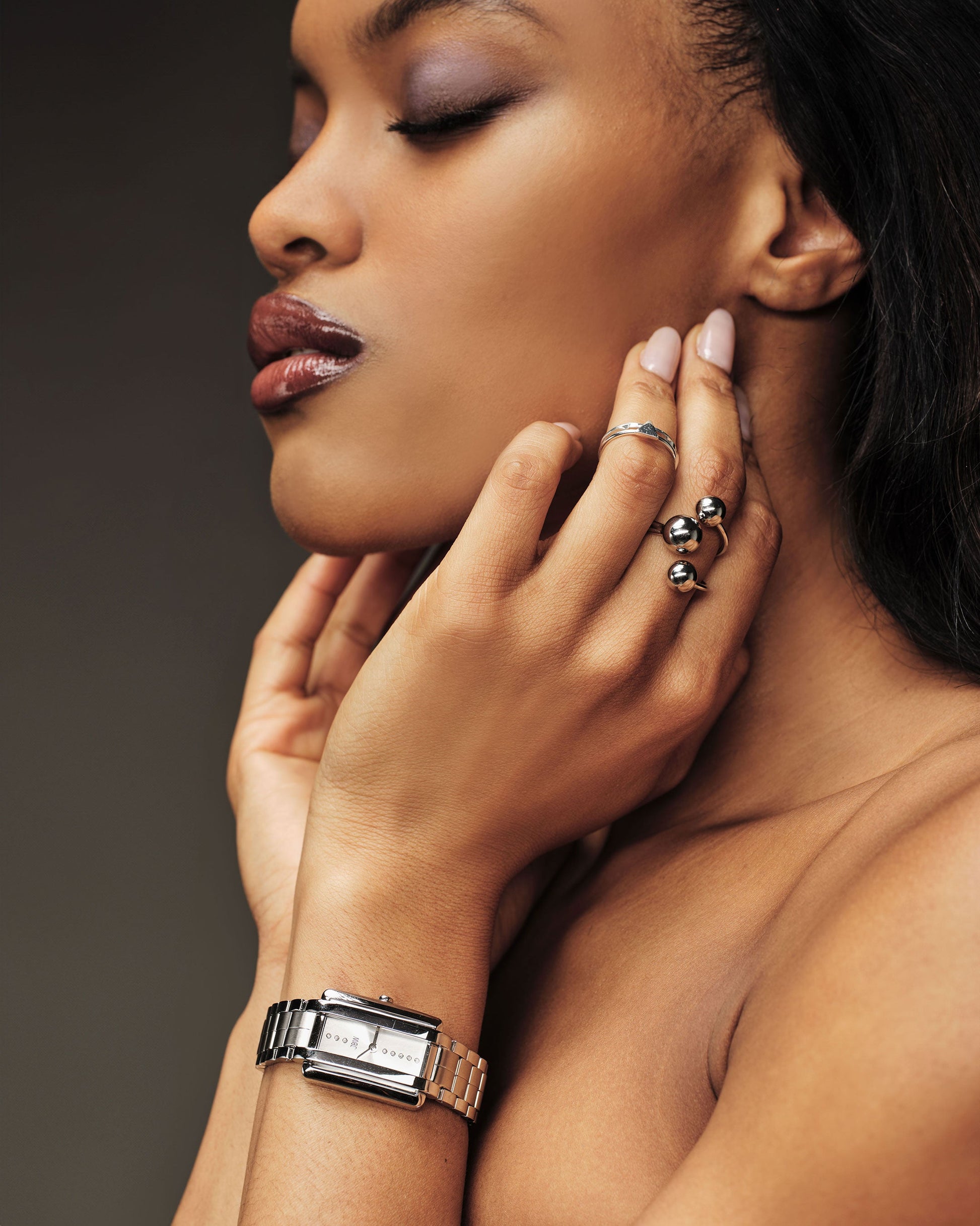 A woman with closed eyes and glossy lips gently touches her neck, showcasing a JBW Watches luxury watch and Mink Petite | J6395B-GA Sample Sale (Grade A) rings with lab grown diamonds on her fingers against a soft brown background.