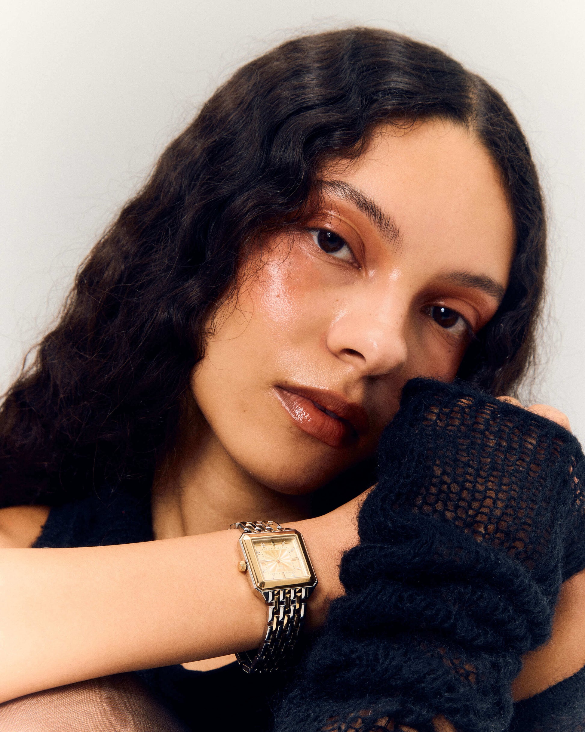 A woman with wavy brown hair and natural makeup gazes at the camera, resting her chin on her arm. She wears the JBW Watches Astra | J6410C square watch and black, loosely knit fingerless gloves.