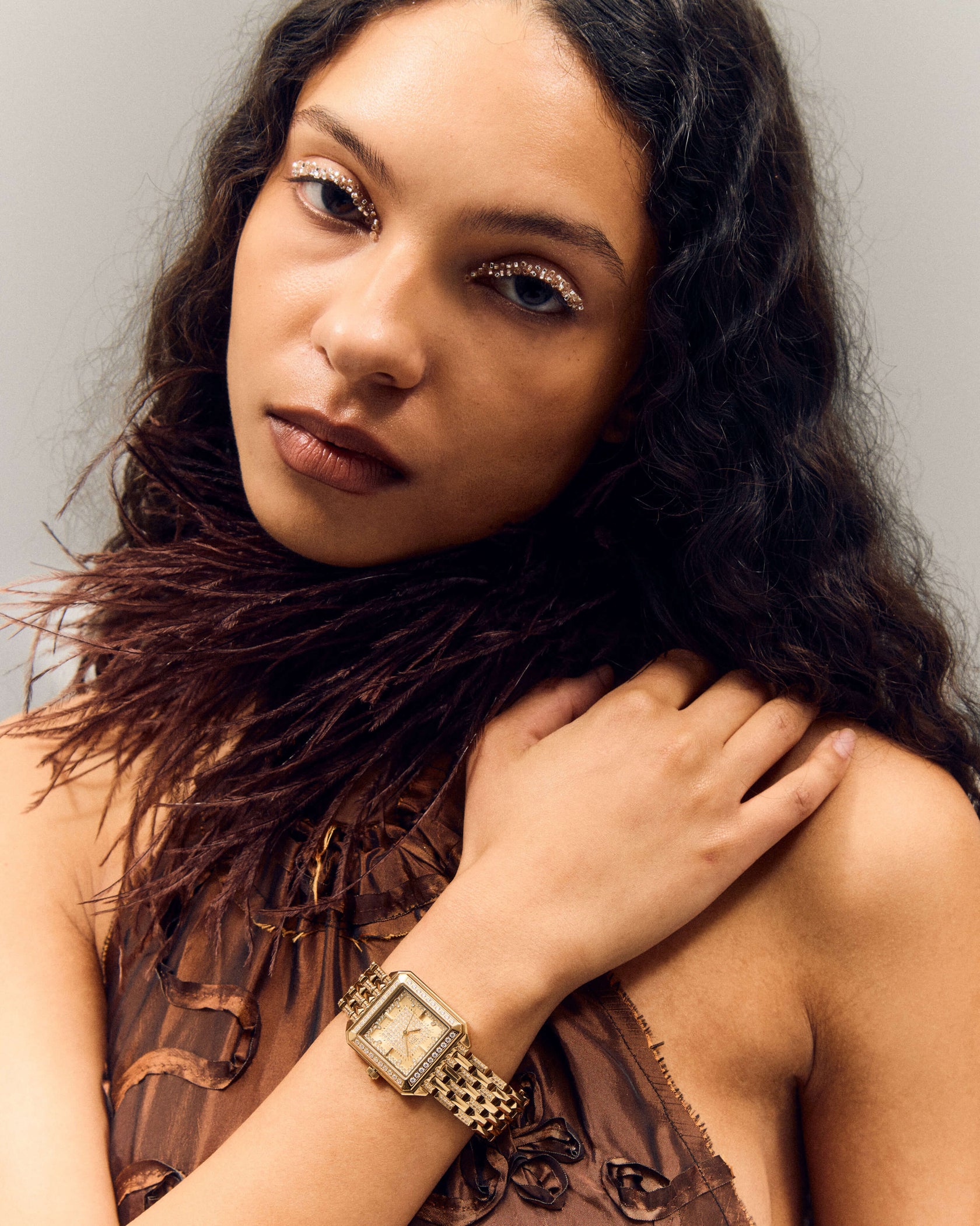 A woman with long, wavy hair and glittery eye makeup gazes at the camera, wearing a brown feathered top and the JBW Watches Astra | J6411B—a square silhouette watch with a pavé-grid dial—resting her hand on her shoulder.