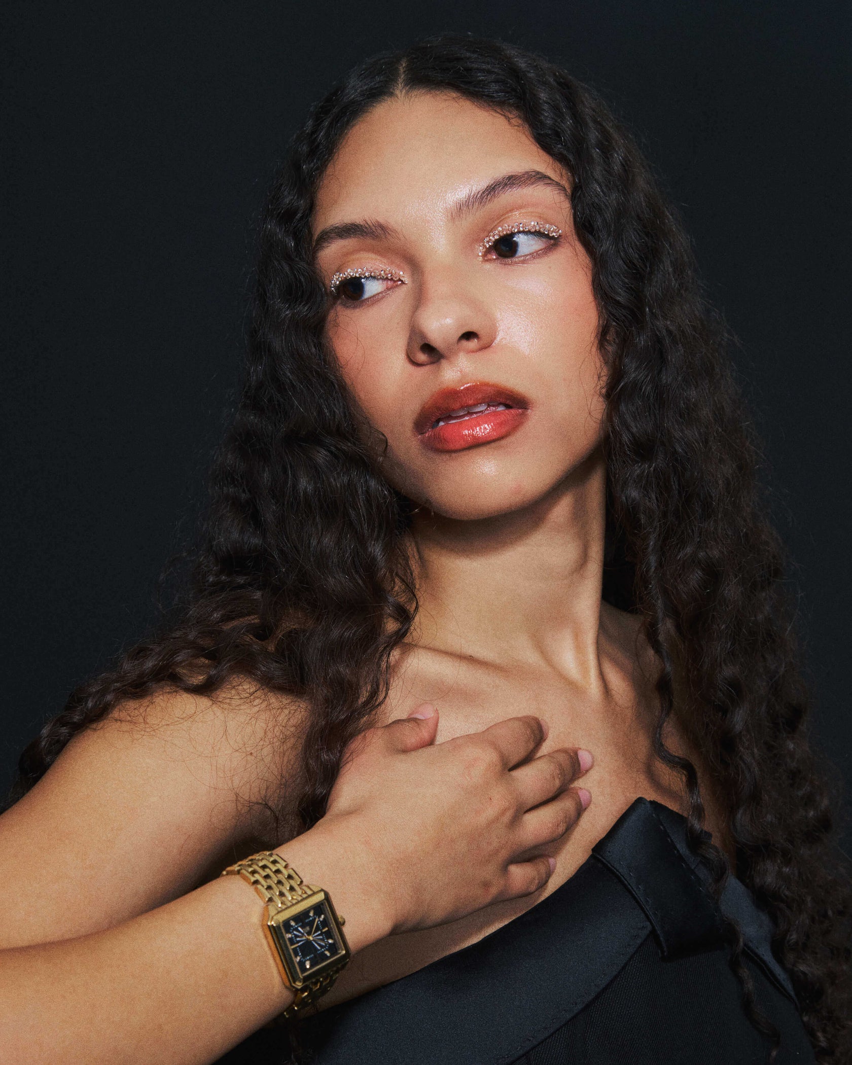 A woman with long curly hair and glossy makeup looks to the side, wearing a black strapless top and the JBW Watches Astra | J6410D—a stainless steel timepiece with a guilloché-textured dial—her hand resting on her chest against a dark background.