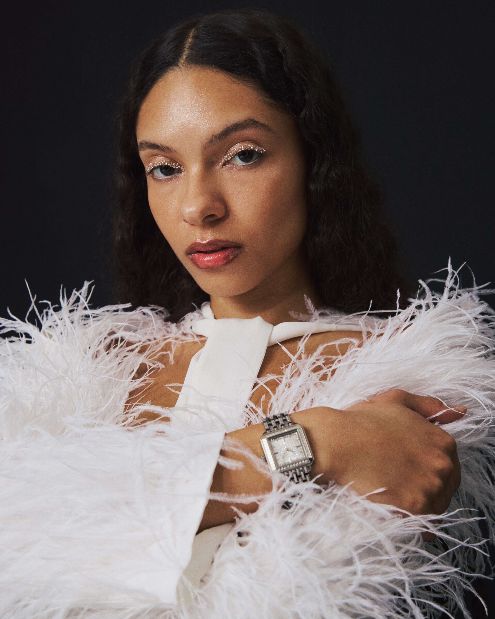 A woman with wavy dark hair wears a white feathered outfit and the square silhouette Astra | J6411A watch by JBW Watches, posing with one arm crossed over her chest against a dark background. Her makeup features shimmery eyeshadow and glossy lips.