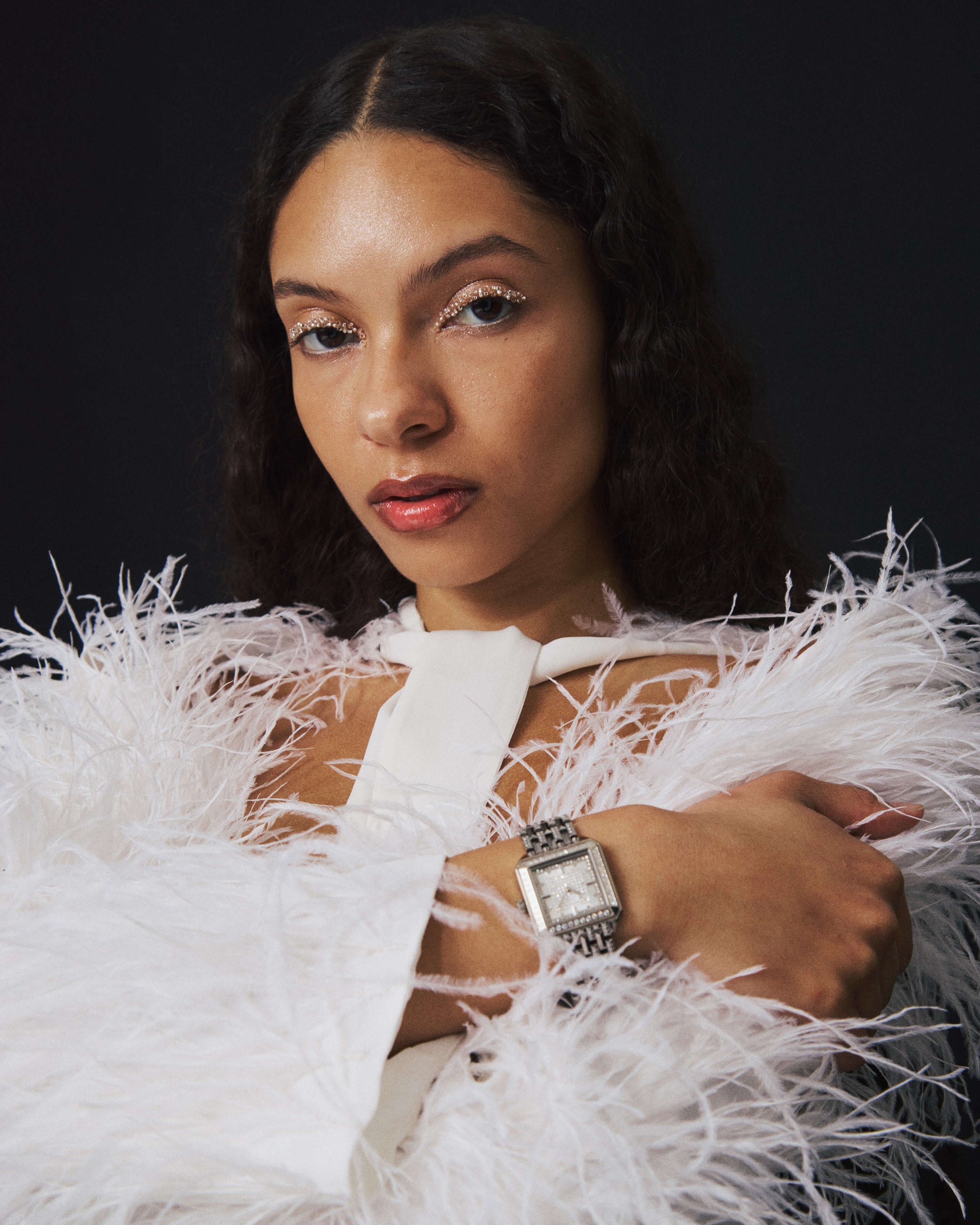 A woman with wavy dark hair wears a white feathered outfit and the square silhouette Astra | J6411A watch by JBW Watches, posing with one arm crossed over her chest against a dark background. Her makeup features shimmery eyeshadow and glossy lips.