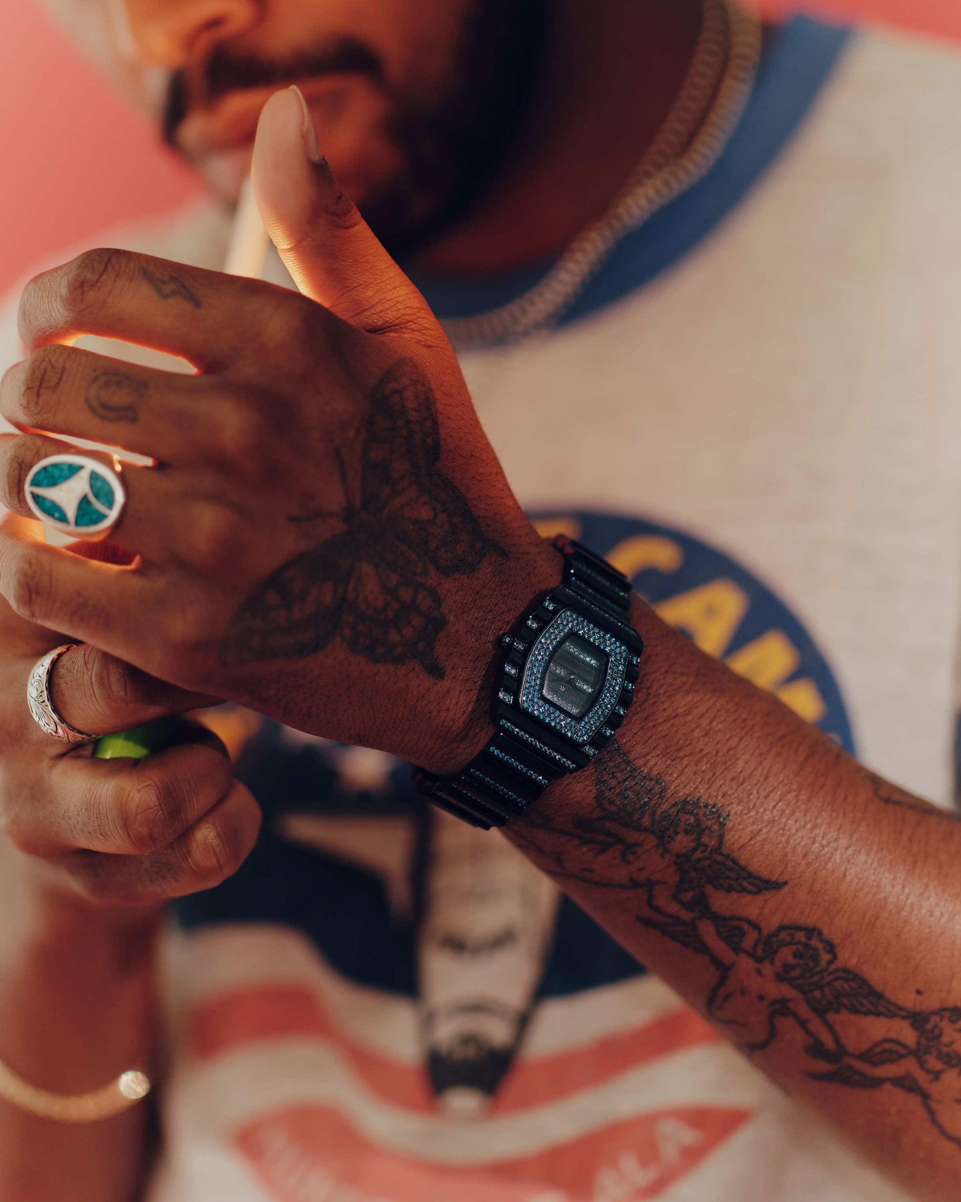 A tattooed individual with a peace sign ring lights a cigarette, sporting the limited edition JBW Watches RESERVE Atlas Bleu | R6404A blue crystal Swiss quartz watch and a vibrant graphic tee. The focus is on their hands and accessories.