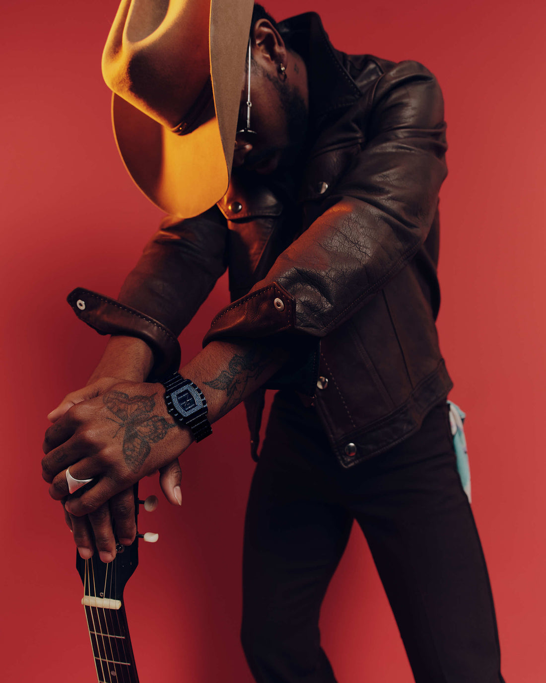 A person in a brown leather jacket and wide-brimmed hat leans forward, resting tattooed arms—one wearing the JBW Watches RESERVE Atlas Bleu | R6404A—on a guitar neck, set against a solid red background.