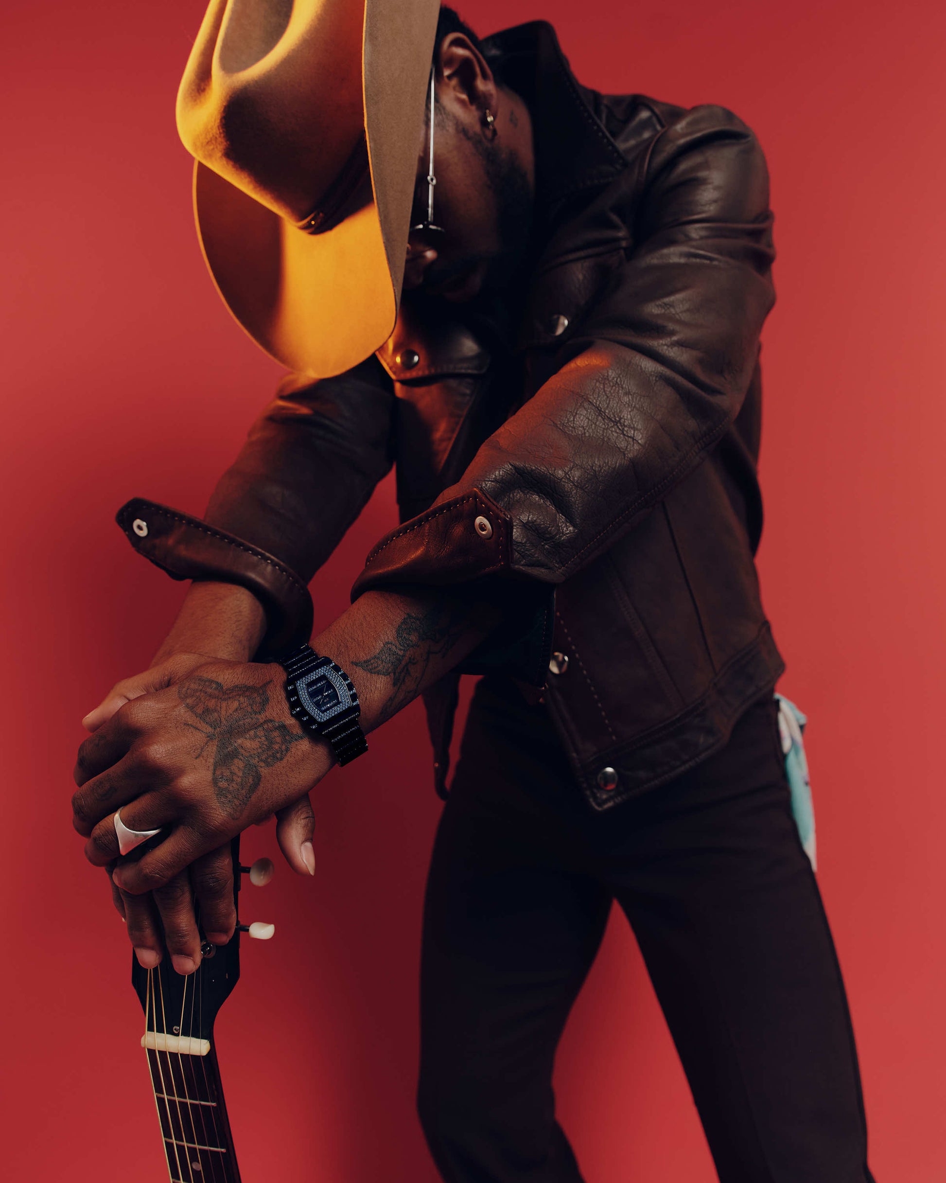 A person in a brown leather jacket and wide-brimmed hat leans forward, resting tattooed arms—one wearing the JBW Watches RESERVE Atlas Bleu | R6404A—on a guitar neck, set against a solid red background.