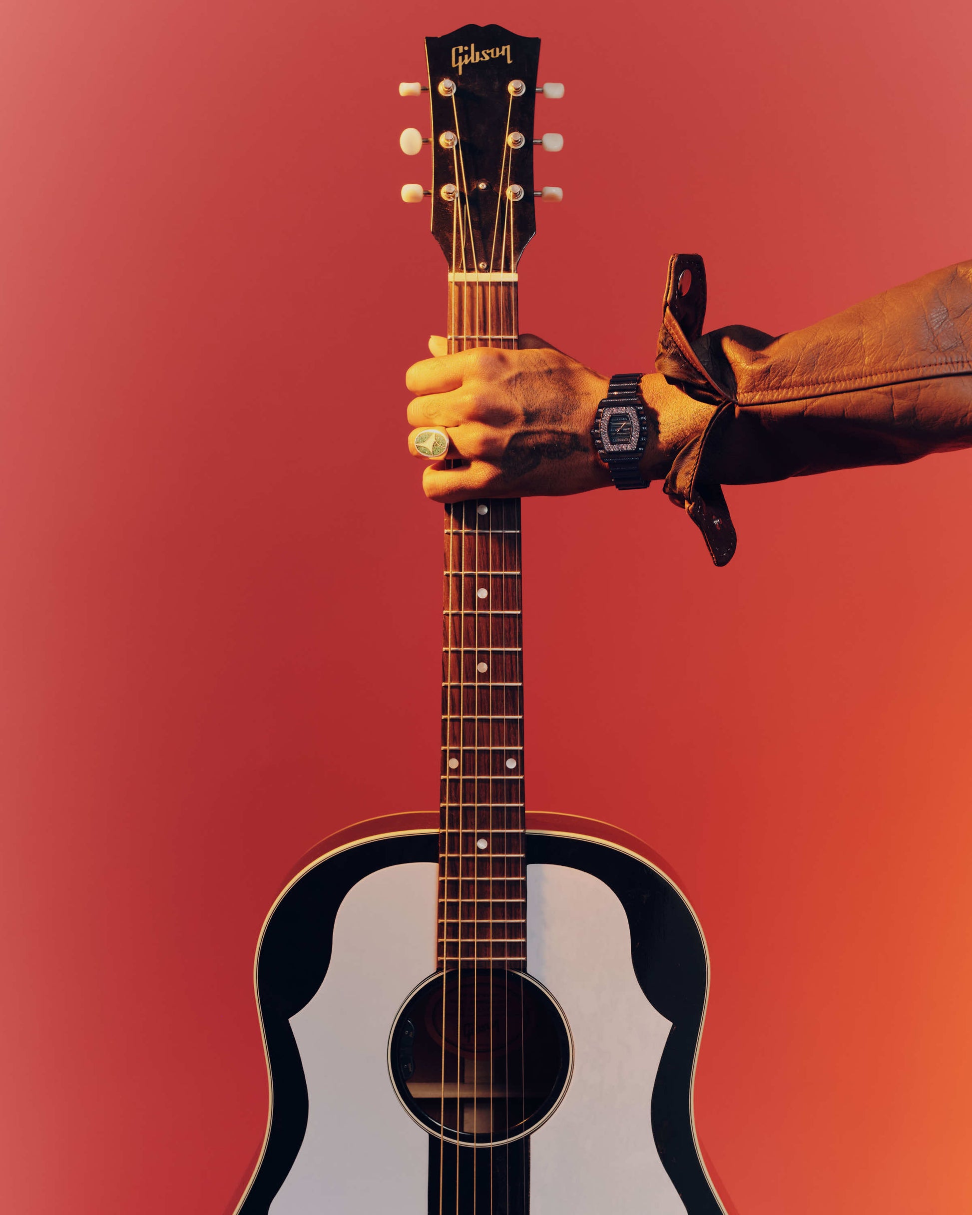 A person wearing a brown jacket and the JBW Watches RESERVE Atlas Bleu | R6404A grips the neck of an acoustic Gibson guitar against a red-orange background.
