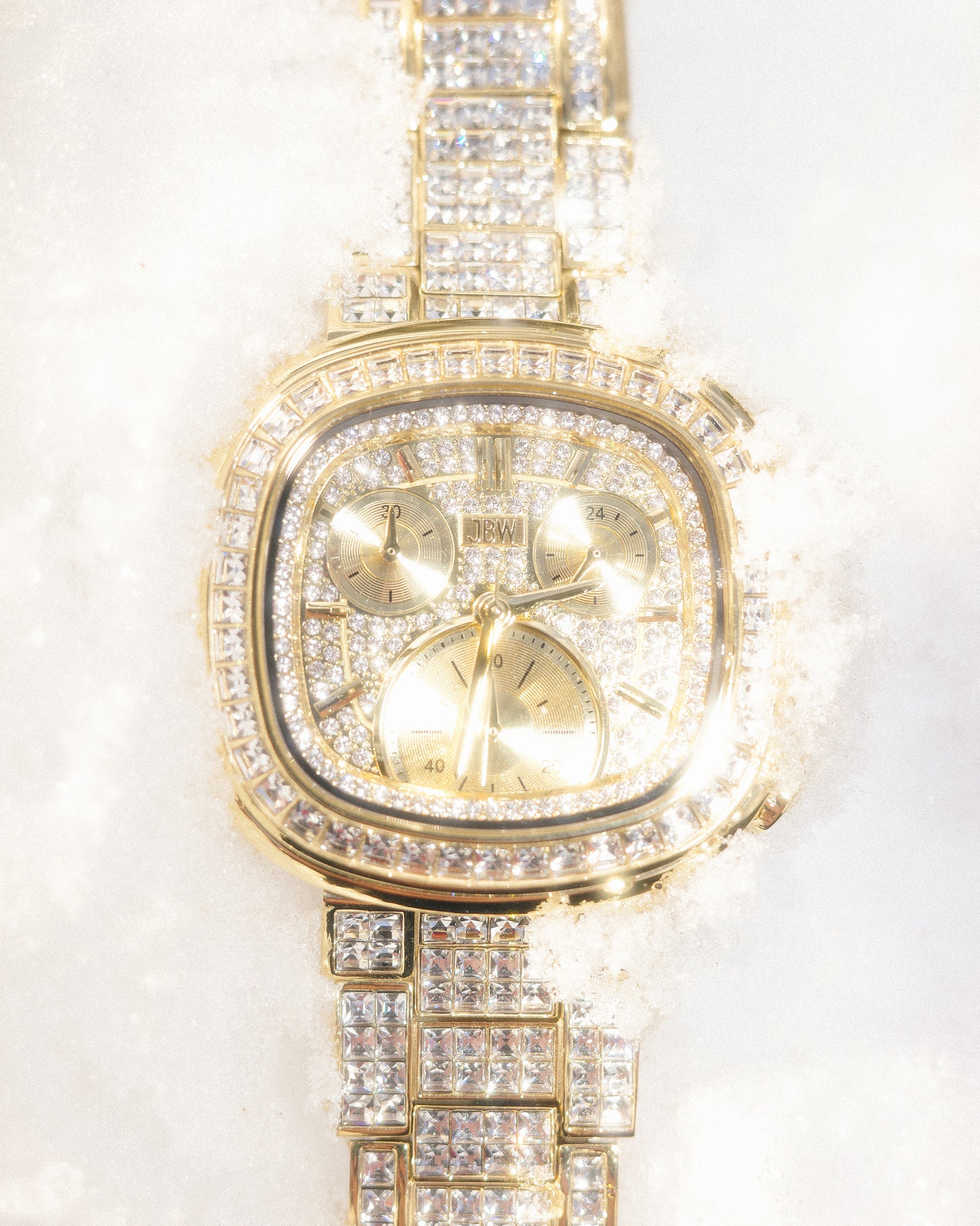 The JBW Watches RESERVE Coast Chandelier | R6397D, a gold chronograph with lab-grown diamonds and three subdials, is shown partially embedded in white snow, its gold-tone bracelet sparkling in the bright light.