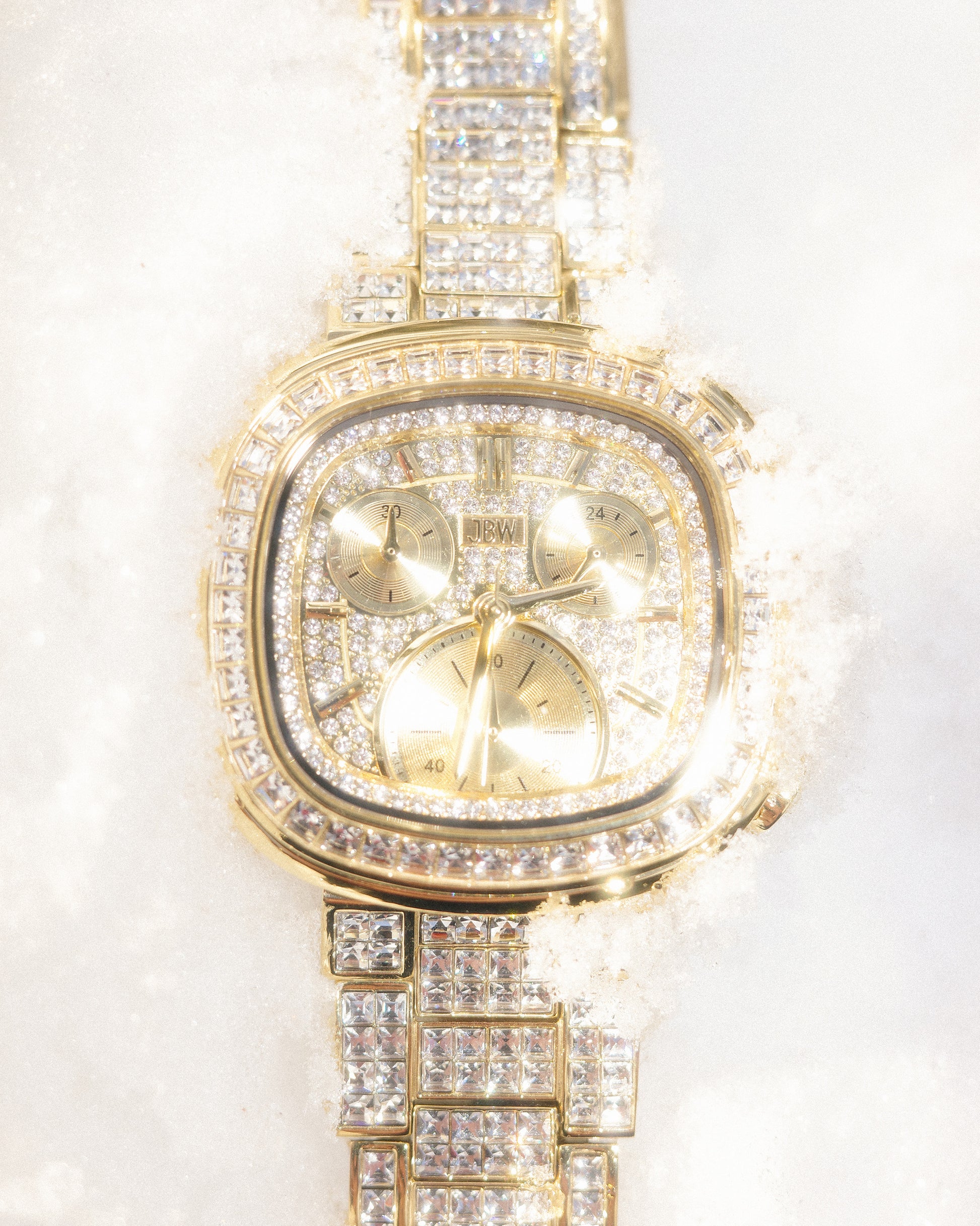 The JBW Watches RESERVE Coast Chandelier | R6397D, a gold chronograph with lab-grown diamonds and three subdials, is shown partially embedded in white snow, its gold-tone bracelet sparkling in the bright light.