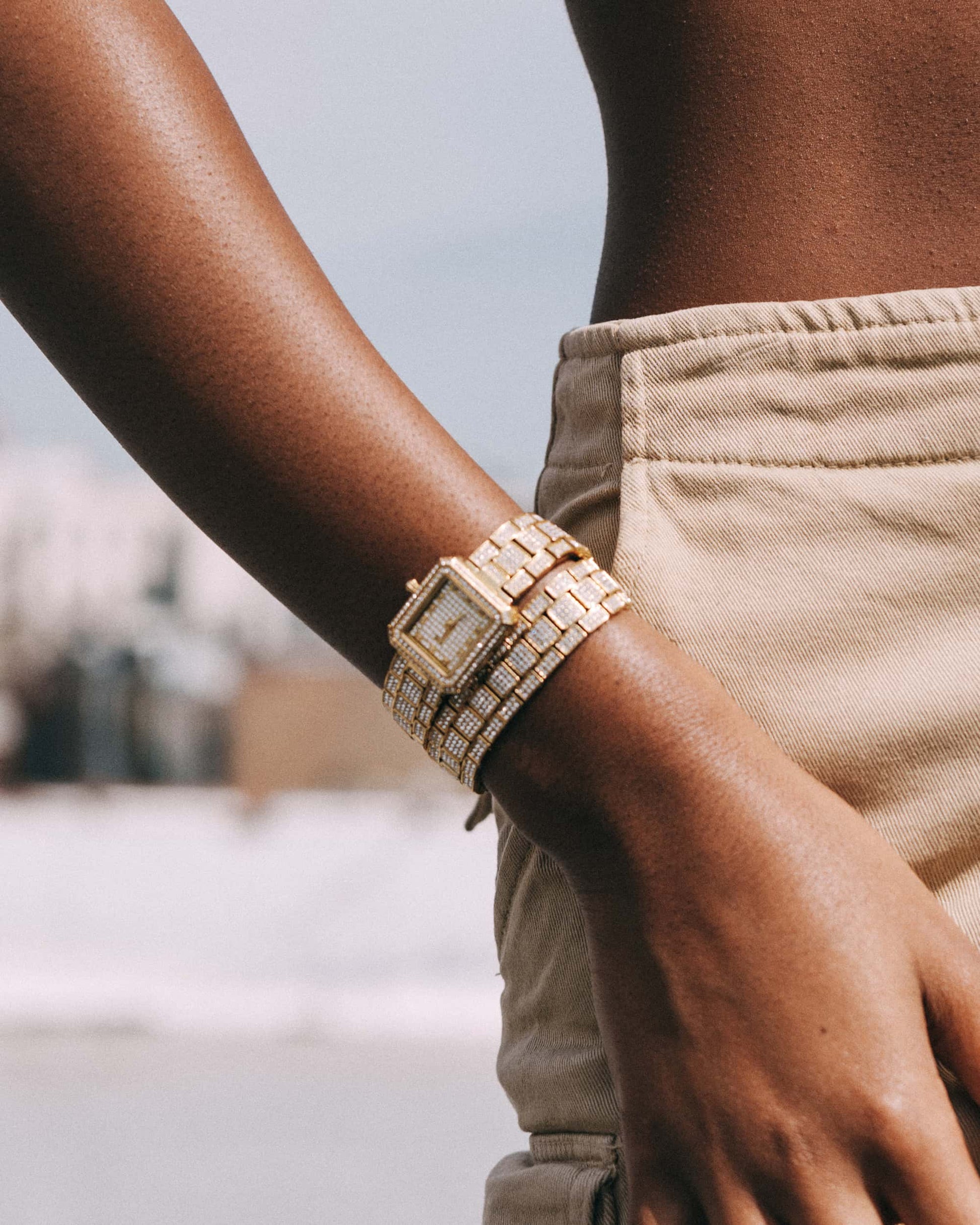 A close-up of a person wearing beige pants and the JBW Watches Arc Double (Sample Sale) | J6390C-GA, featuring a crystal-studded bezel, with a blurred outdoor background.