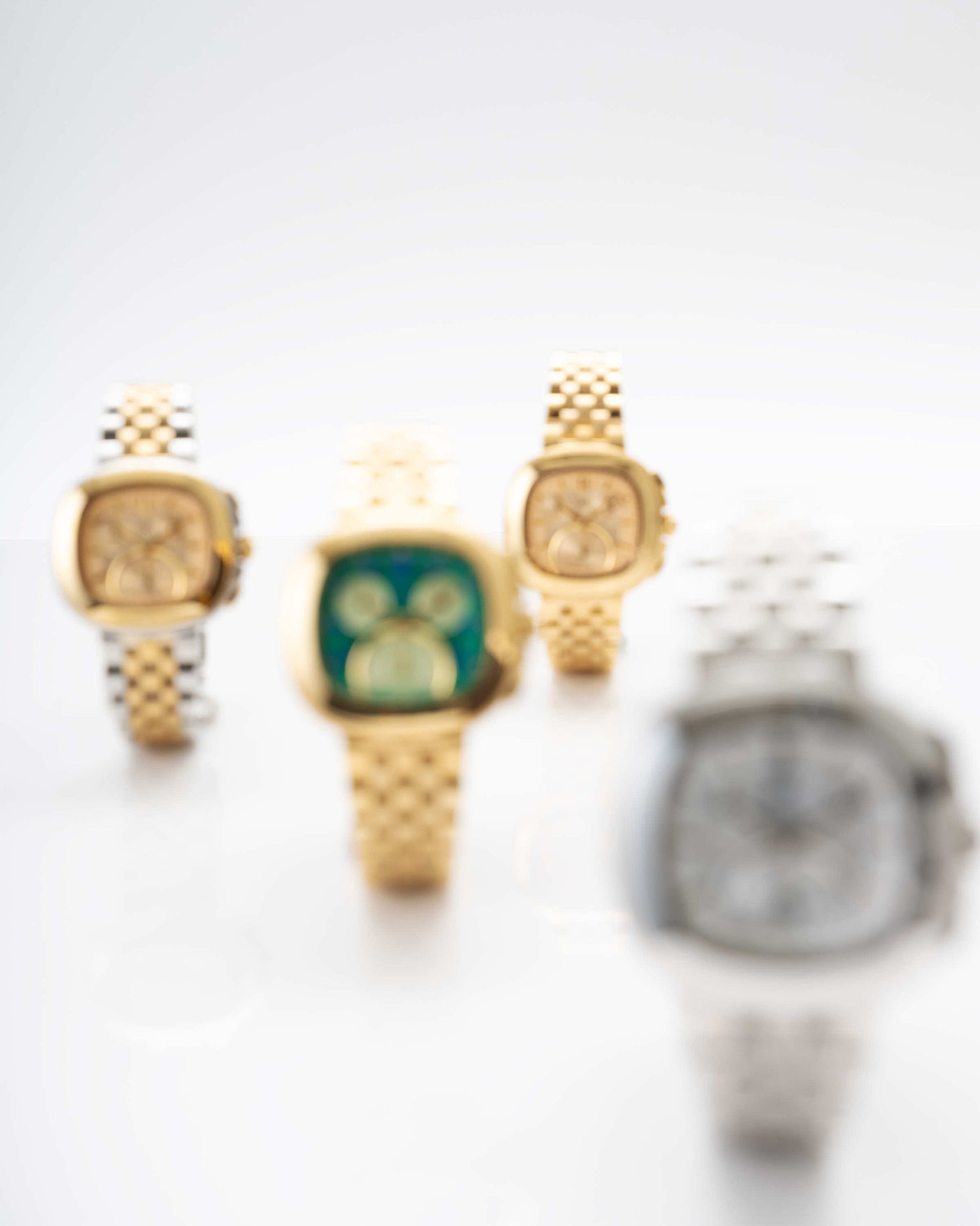 Four JBW Watches are shown on a light surface, featuring the Coast | J6397A model. Three gold watches are in the background and one stainless steel watch is in the front. All watches, possibly including a chronograph, are out of focus.