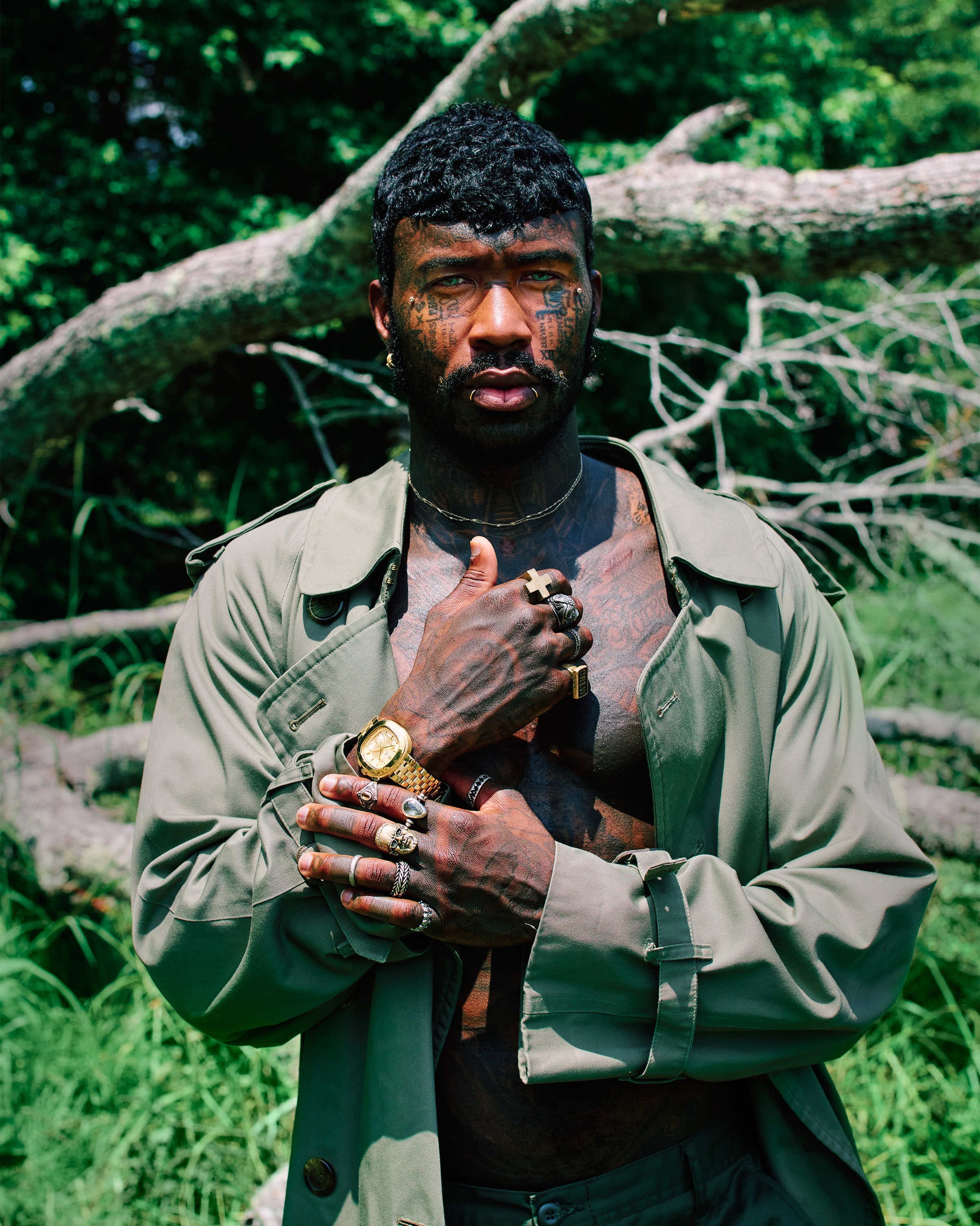 A man with facial tattoos and rings poses outdoors in front of a fallen tree, wearing an open green trench coat and showcasing the JBW Watches Coast | J6397A stainless steel watch amidst lush greenery.
