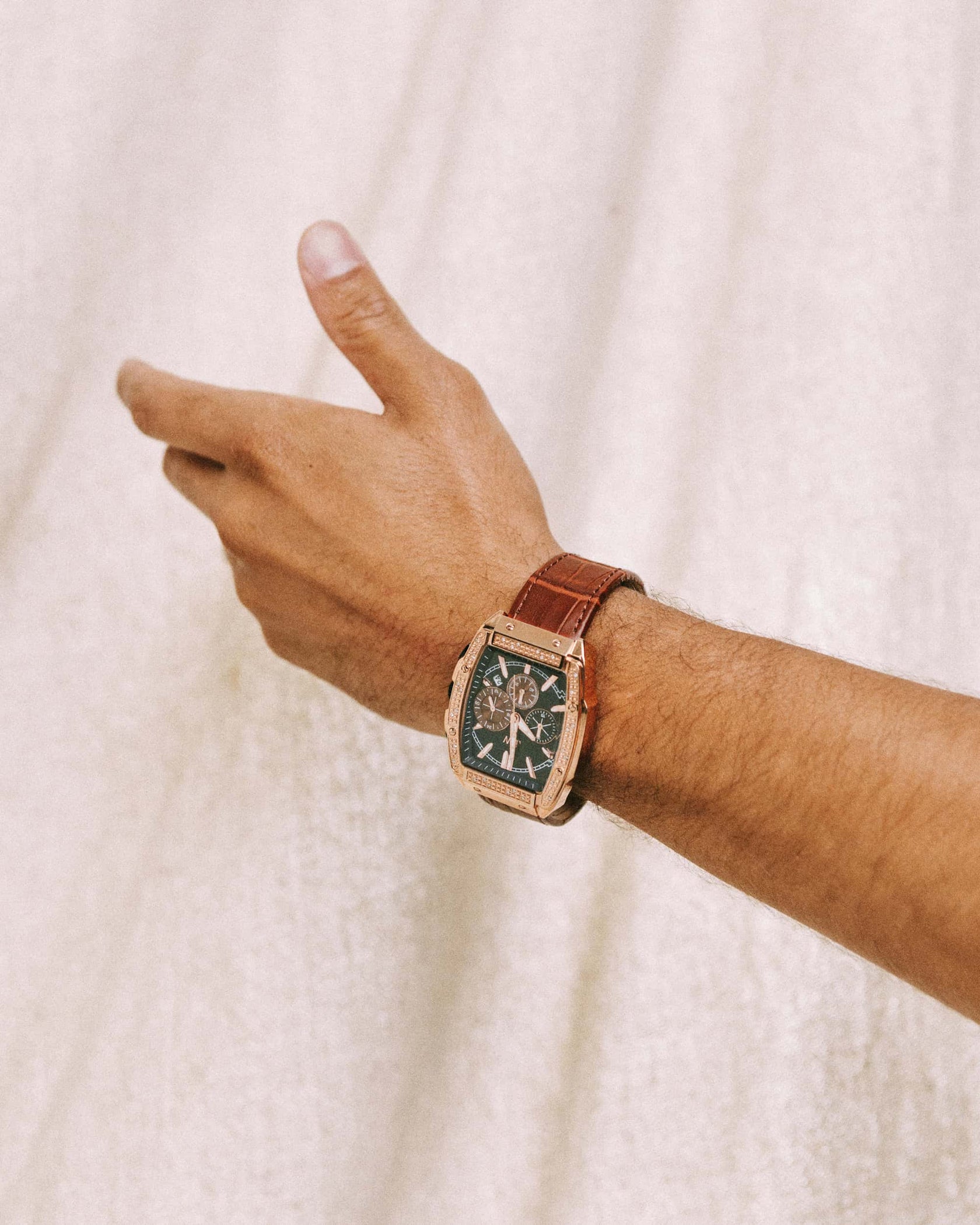 A hand with light brown skin wears the JBW Echelon | J6379D, a diamond chronograph watch with a rectangular case and embossed leather band, set against a softly textured light background.