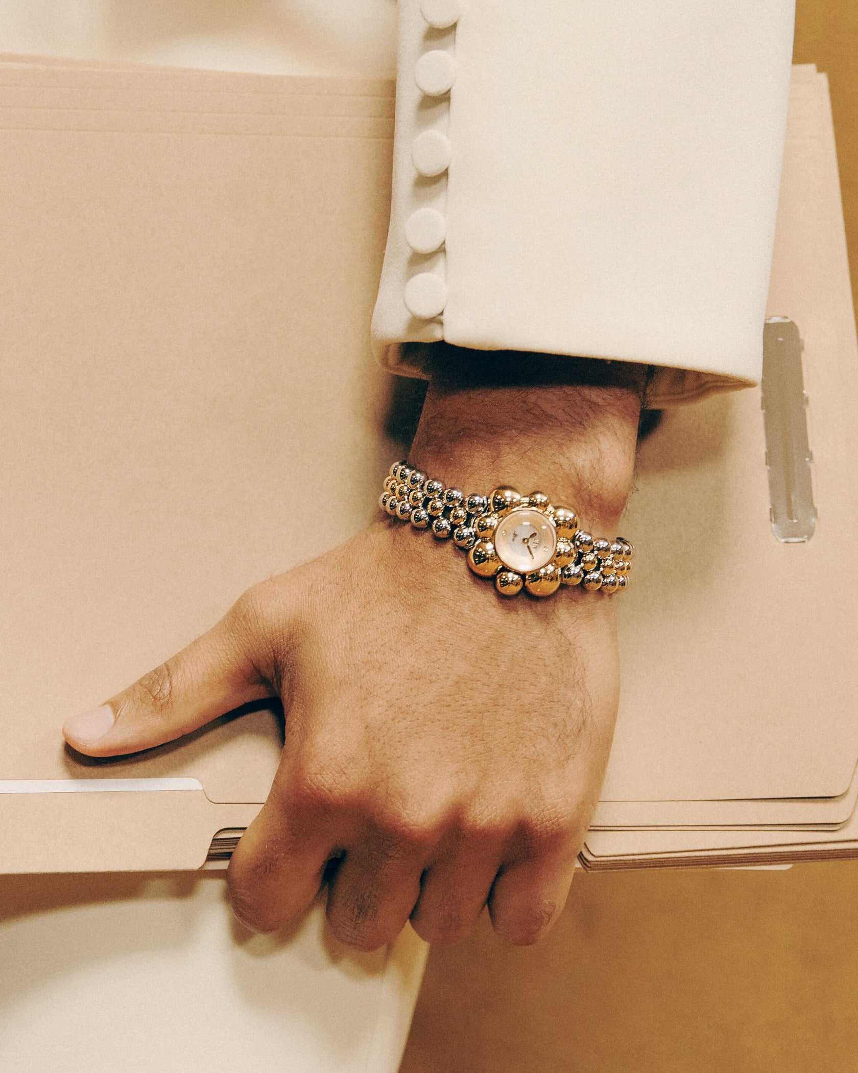 A person in a cream suit with fabric-covered buttons holds a beige file folder under their arm, wearing the JBW x Luar – Atmosphere (Sample Sale) stainless steel gold watch with a mother of pearl dial from JBW Watches.