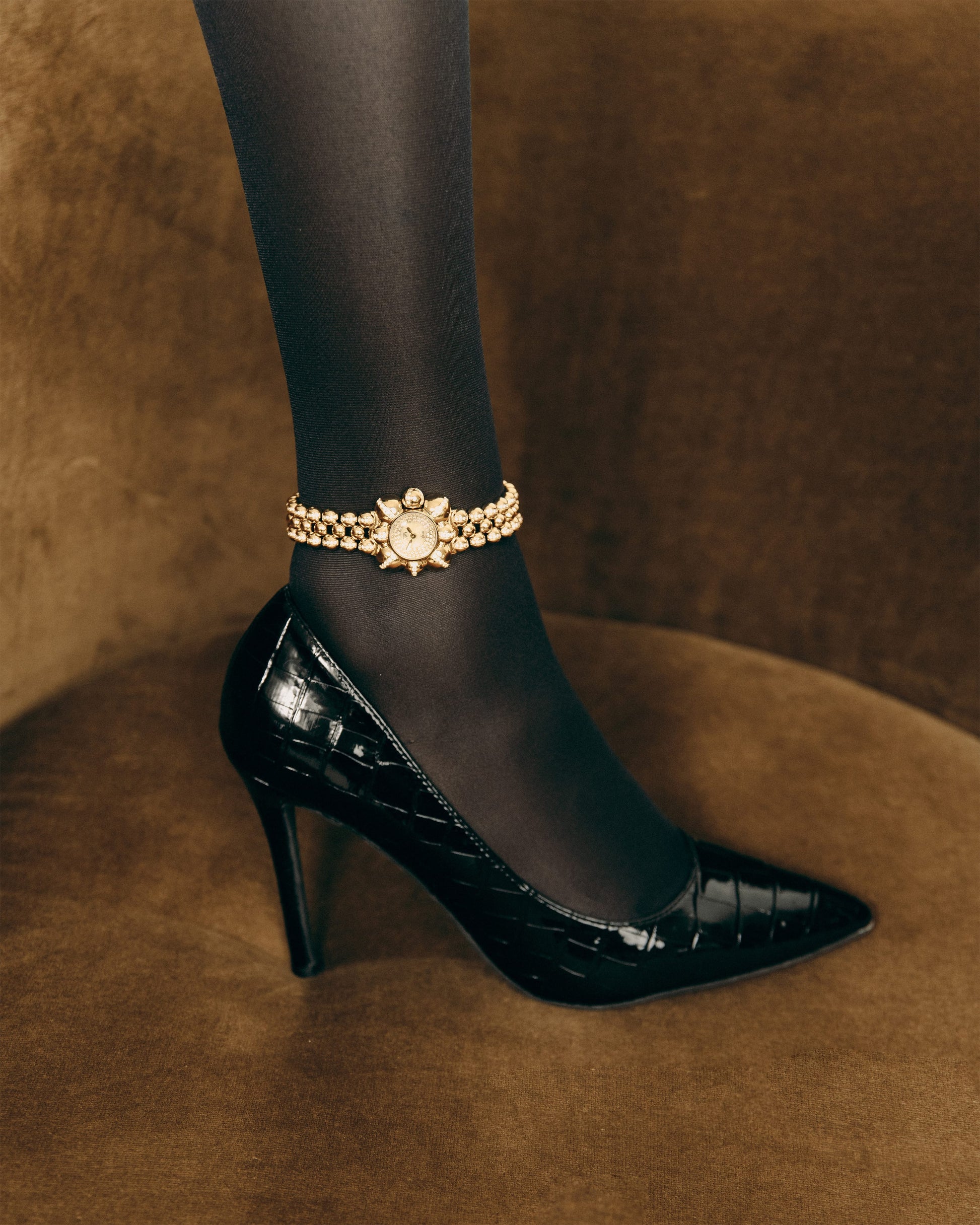 A person wearing black sheer tights, a black patent leather high heel, and a JBW x Luar – Atmosphere (Sample Sale) | J6406A-SC watch by JBW Watches sits on a brown velvet surface.