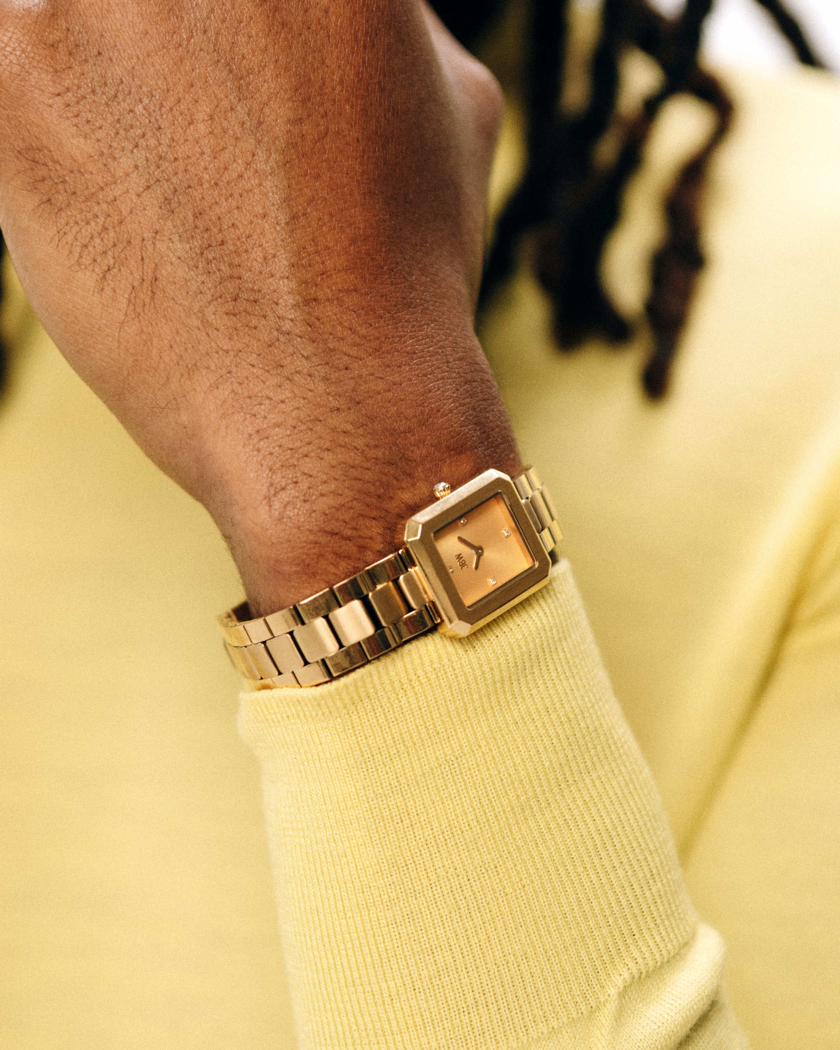 A close-up of a wrist wearing the JBW Watches Arc Single | J6390E, a gold luxury watch with a square face, paired with a yellow long-sleeve shirt. The background is blurred and the wearer has dark, twisted hair.