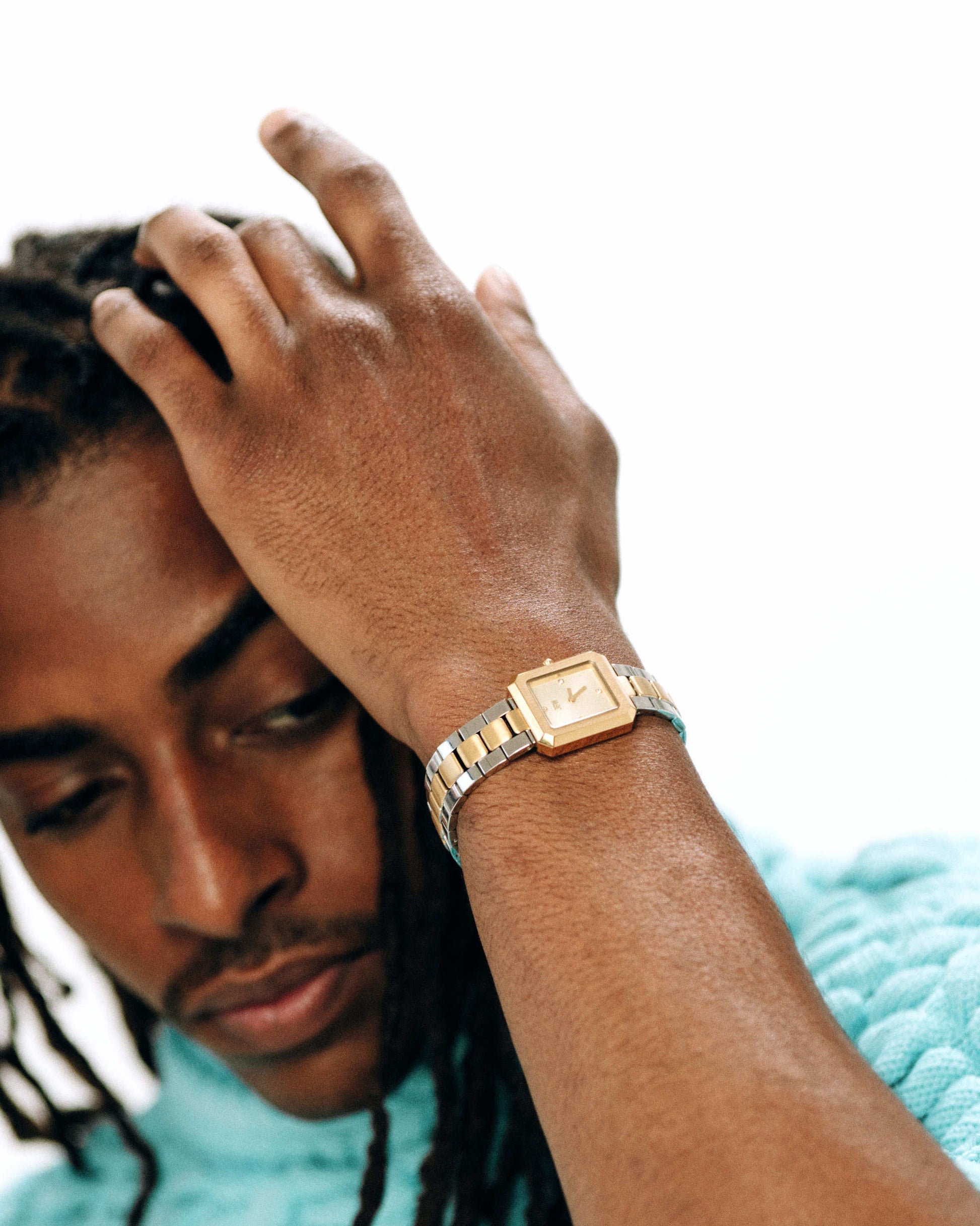 A person with braided hair in a light blue textured sweater raises a hand, revealing the JBW Watches Arc Single | J6390F—a gender-neutral square-face watch with gold and silver accents—against a plain white background.