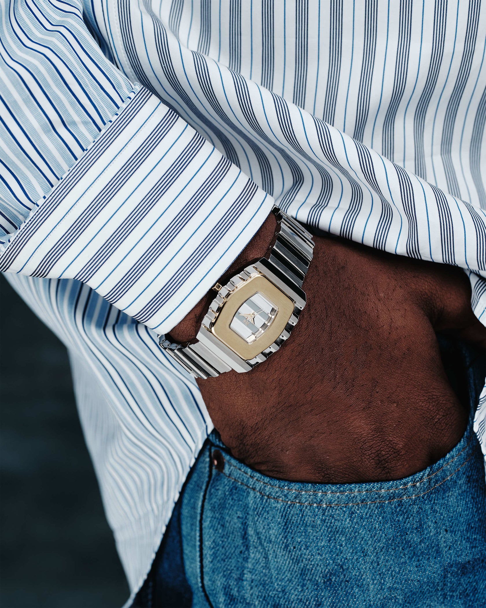 A close-up of a person in a striped shirt and blue jeans, hand in pocket, wearing the JBW Watches Atlas (Sample Sale) | J6403C-SC, featuring a geometric face and stainless steel bracelet.