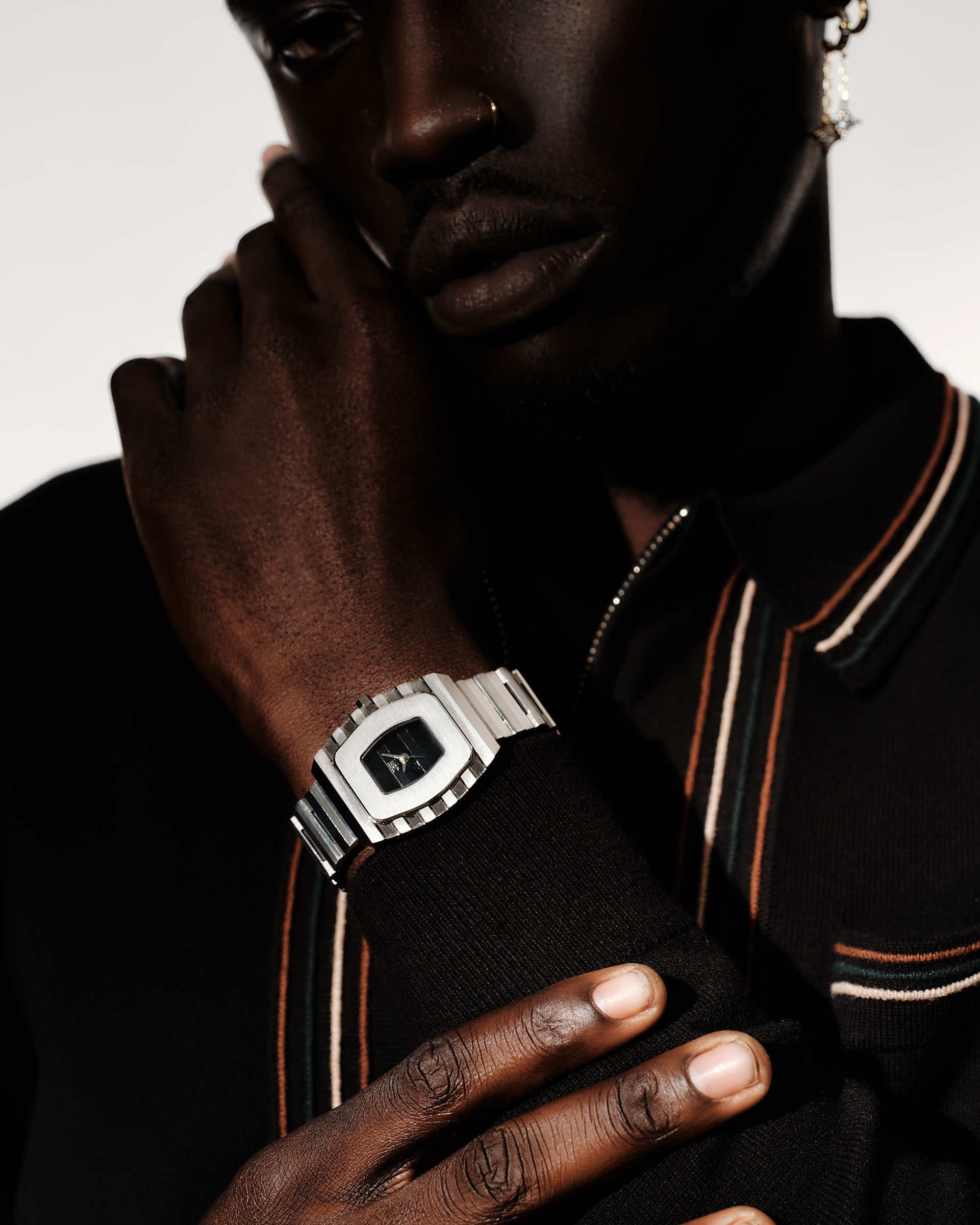 A person with dark skin wears the JBW Watches Atlas (Sample Sale) | J6403D-SC, featuring a stainless steel bracelet, and poses in a black top with brown and white striped trim. Soft lighting highlights the watch and hand as one touches their face.