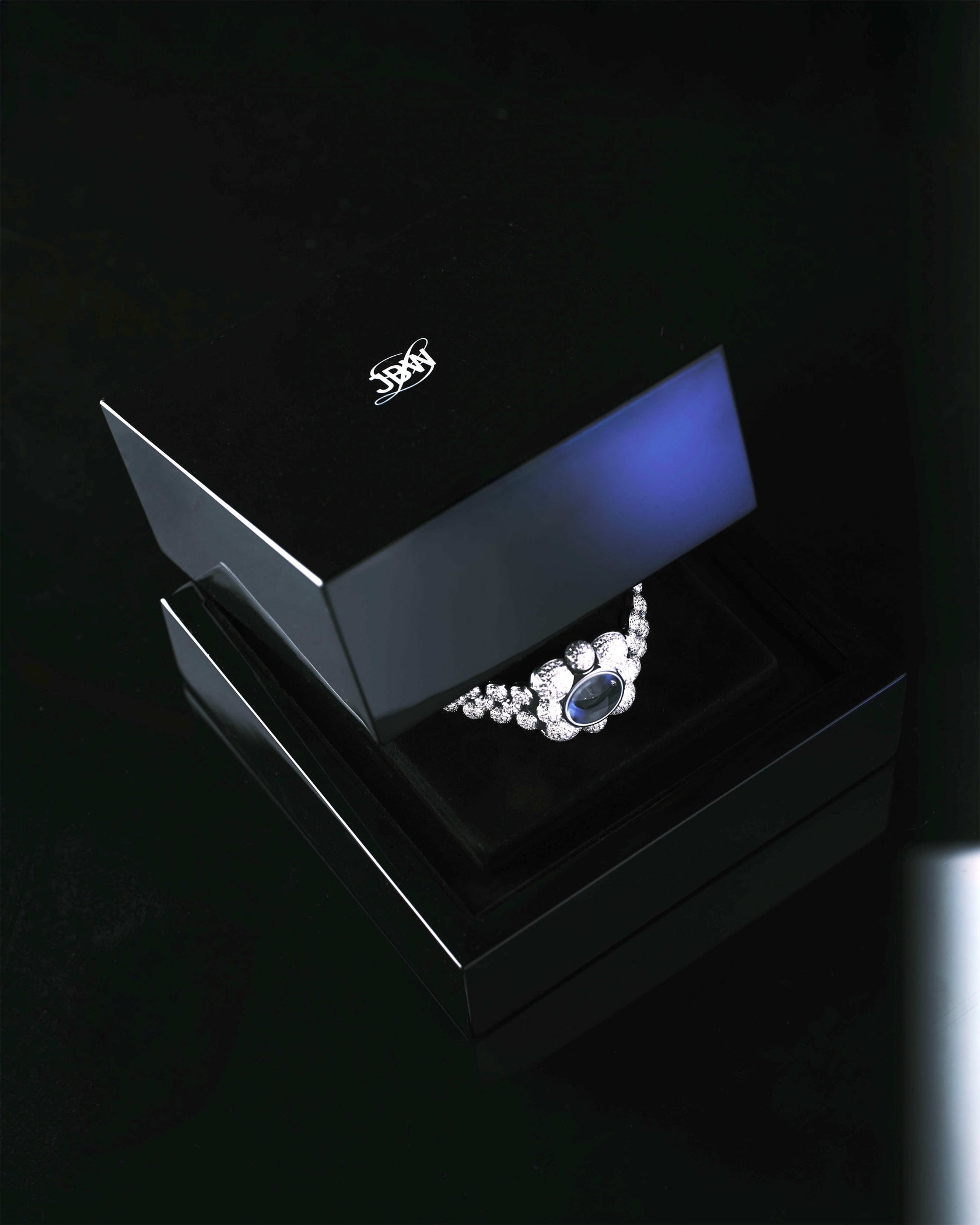 The JBW x Luar – Atmosphere Diamond | J6407A necklace from JBW Watches, featuring a large blue gemstone and lab-grown diamonds, is showcased in an open black jewelry box on a glossy black surface.