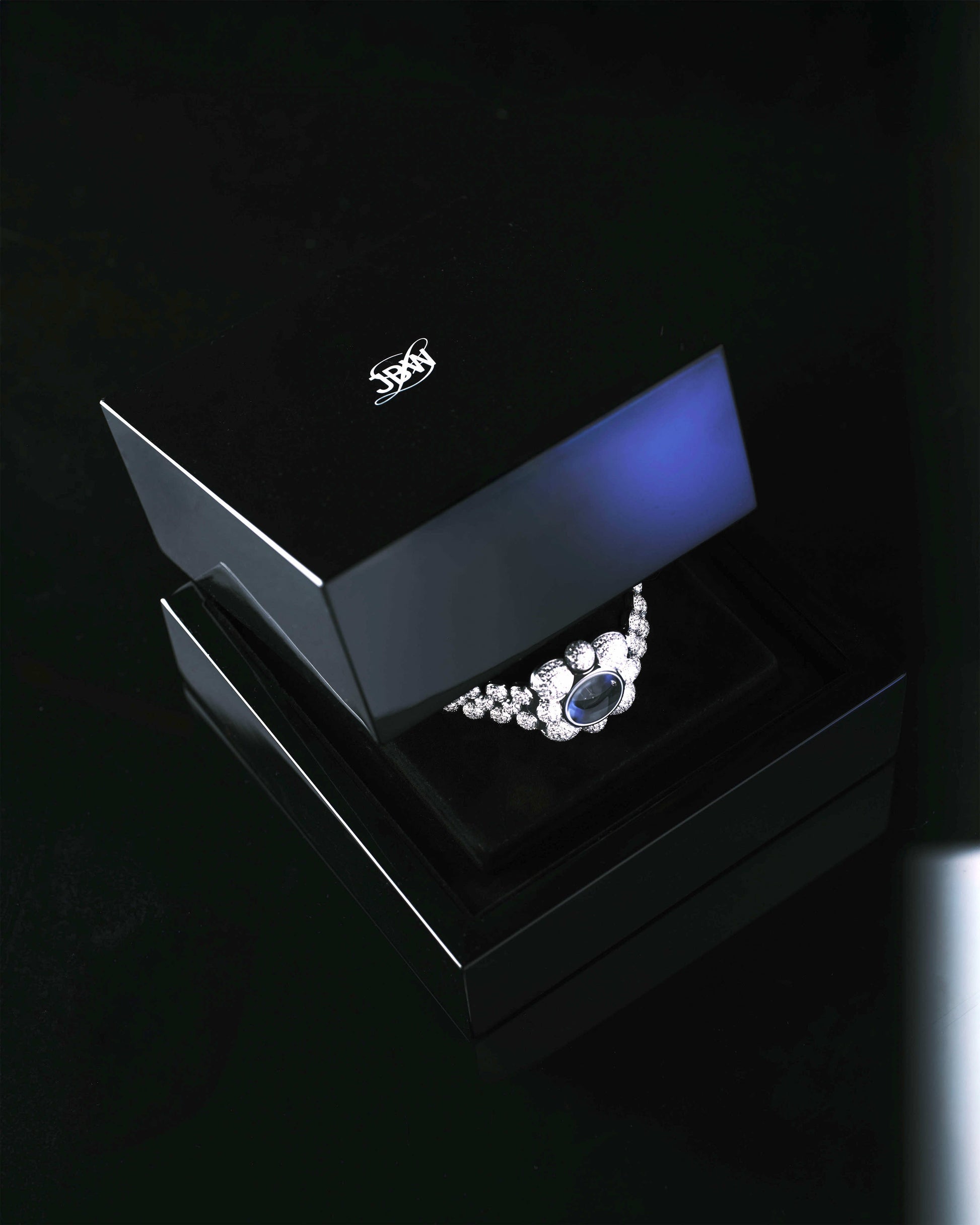 The JBW x Luar – Atmosphere Diamond | J6407A necklace from JBW Watches, featuring a large blue gemstone and lab-grown diamonds, is showcased in an open black jewelry box on a glossy black surface.