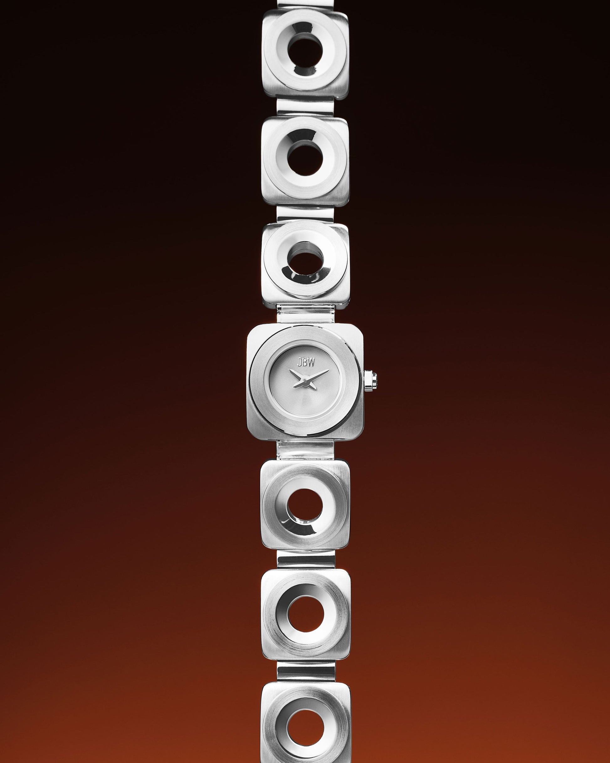 The Chasm Petite (Sample Sale) | J6400B-SC by JBW Watches, a modern silver wristwatch with a square face and geometric links featuring circular cutouts, is showcased vertically on a gradient brown and black background. Japanese VX movement inside.