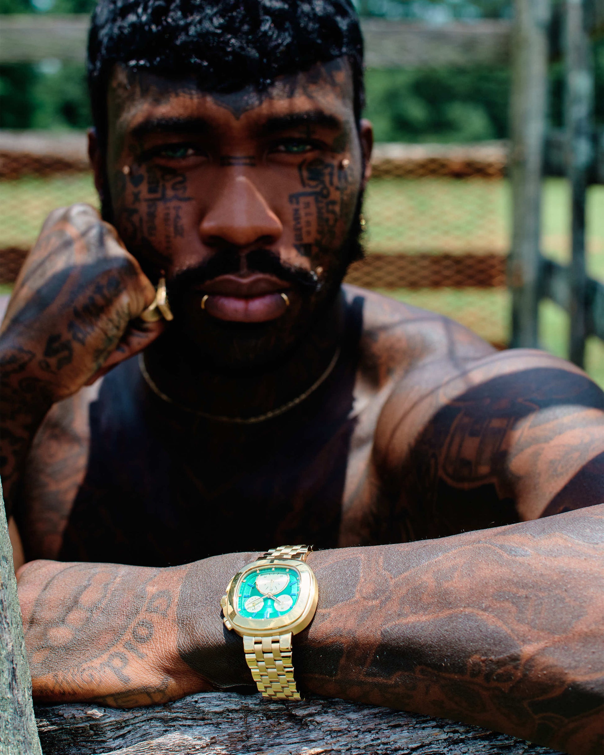A shirtless, tattooed man with facial piercings leans on a wooden fence outdoors, resting his chin on his hand. He wears the JBW Watches Coast | J6397D, a 38mm stainless steel watch with a green face. The background is blurred.