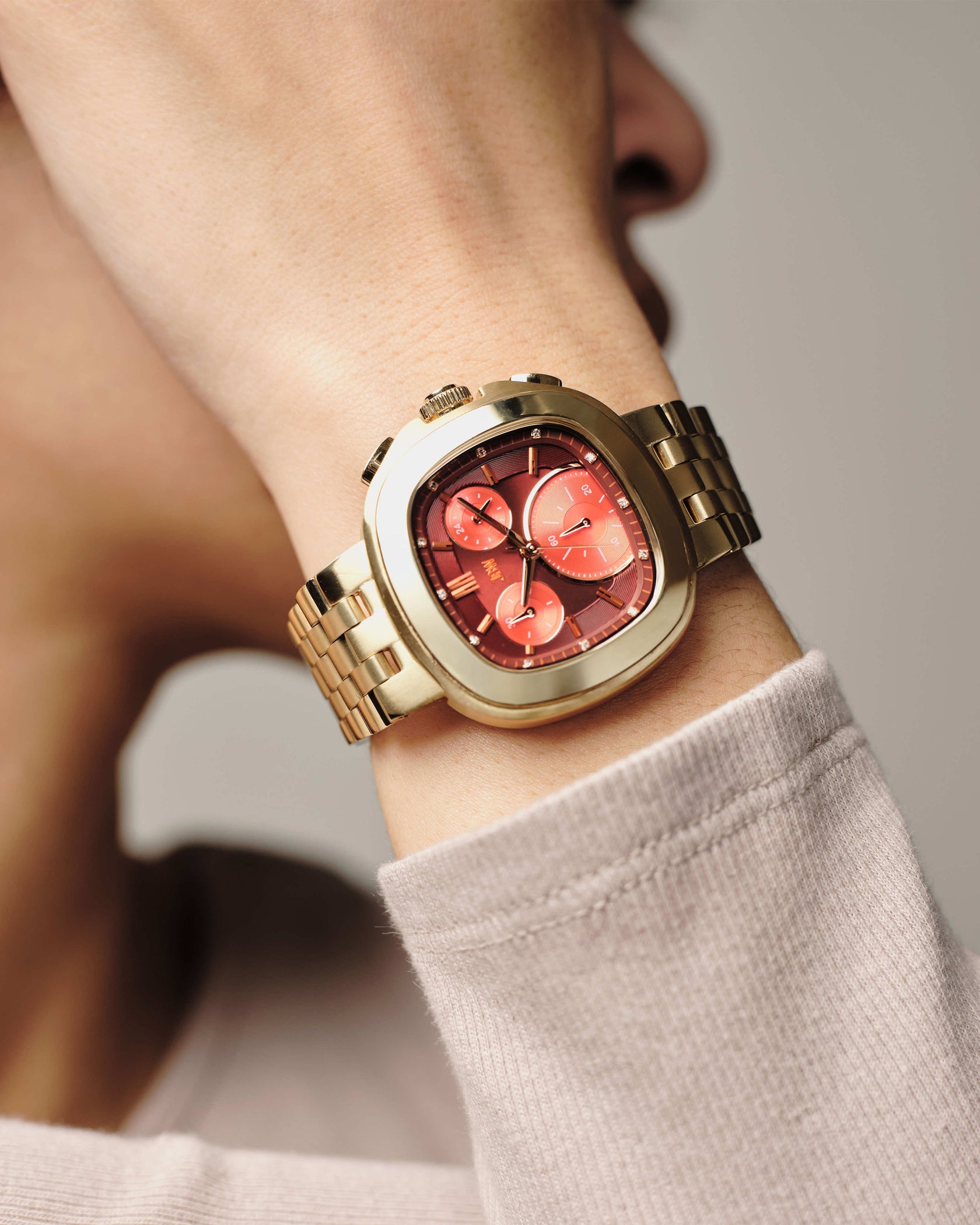 A close-up of a wrist adorned with the JBW Watches Coast | J6397F, a gold watch featuring a red dial with three subdials and Japanese Miyota movement. The wearer sports a light beige long sleeve shirt, hand near their face.