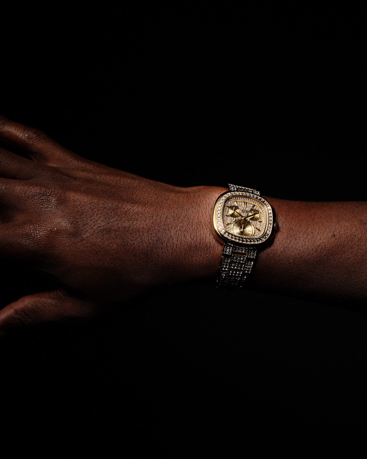 A close-up of a hand and wrist wearing the JBW Watches RESERVE Coast Chandelier | R6397D, an ornate gold-tone bracelet watch with a textured band and detailed face, set against a black background.