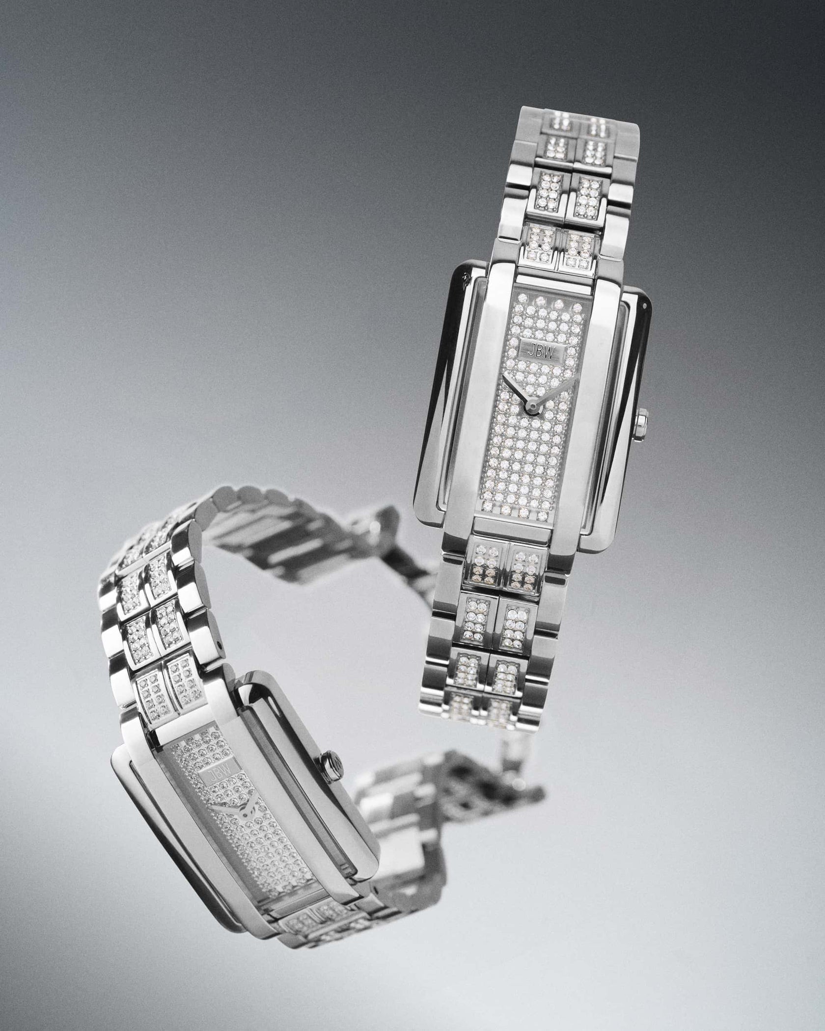 Two luxurious JBW Watches Mink Petite | J6394B silver wristwatches with rectangular faces and lab-grown diamond-encrusted dials and bands are displayed on a gradient gray background, one upright and one lying flat.
