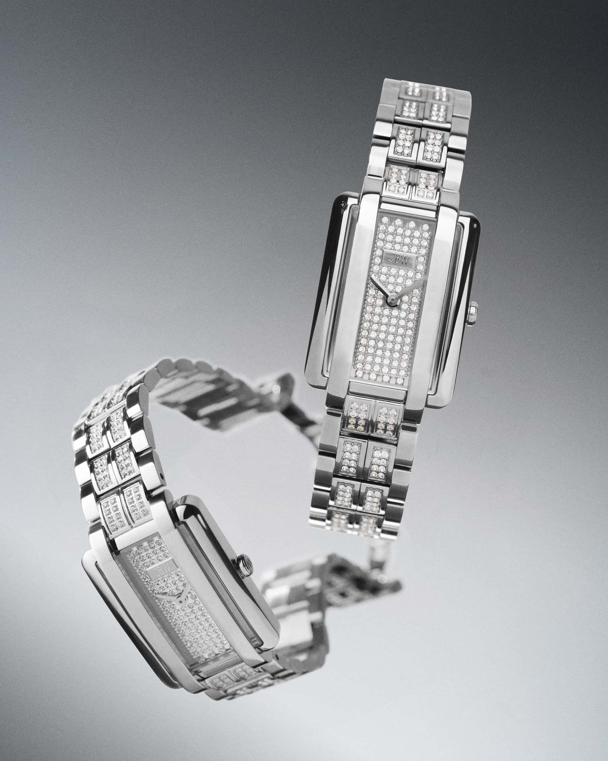 Two luxurious JBW Watches Mink Petite | J6394B silver wristwatches with rectangular faces and lab-grown diamond-encrusted dials and bands are displayed on a gradient gray background, one upright and one lying flat.