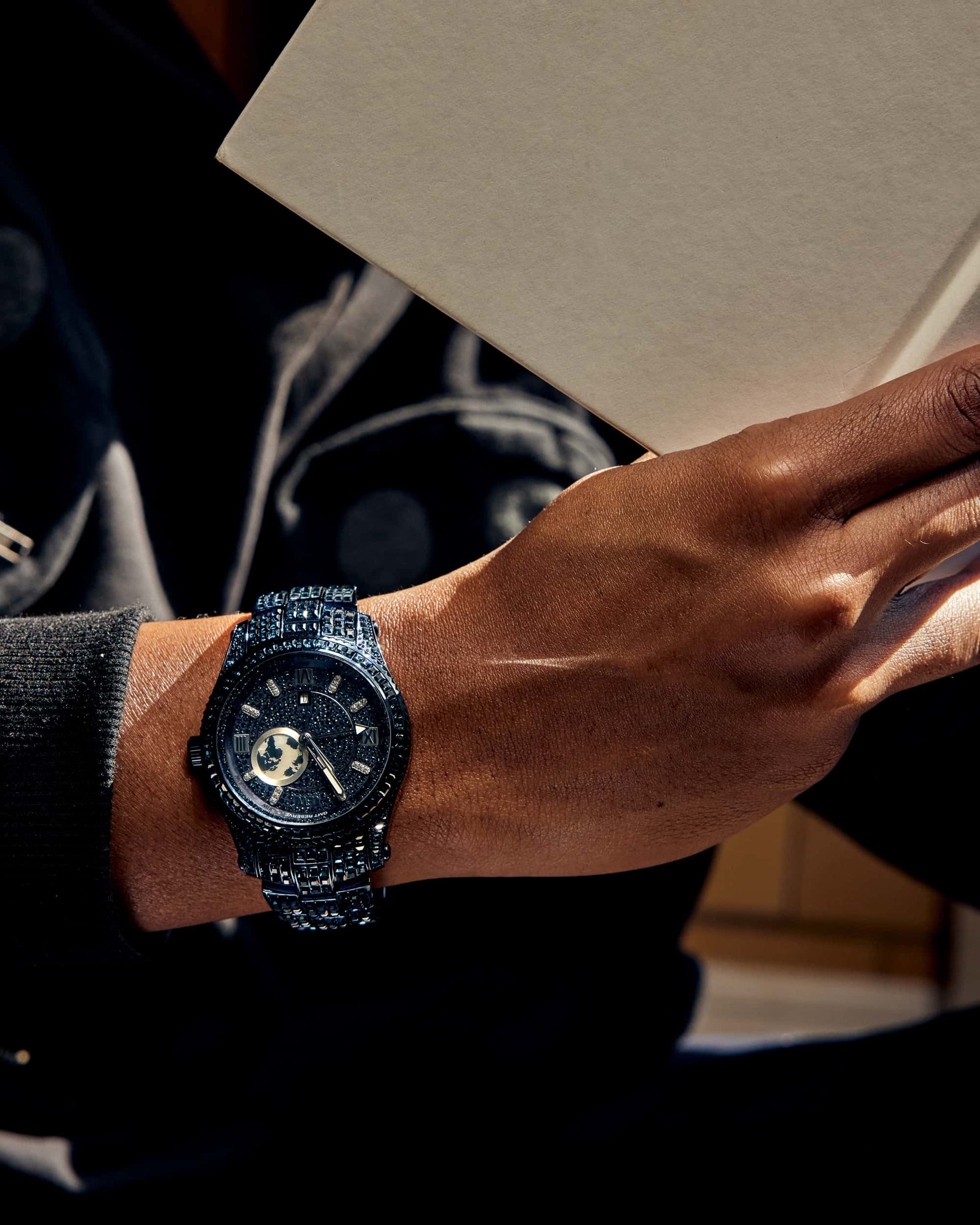 A person in a dark outfit holds an open book, showcasing the RESERVE Bleu 39 | R6393A-GA (Grade A) by JBW Watches. The sapphire blue crystals of the watch catch the eye against a softly blurred background.