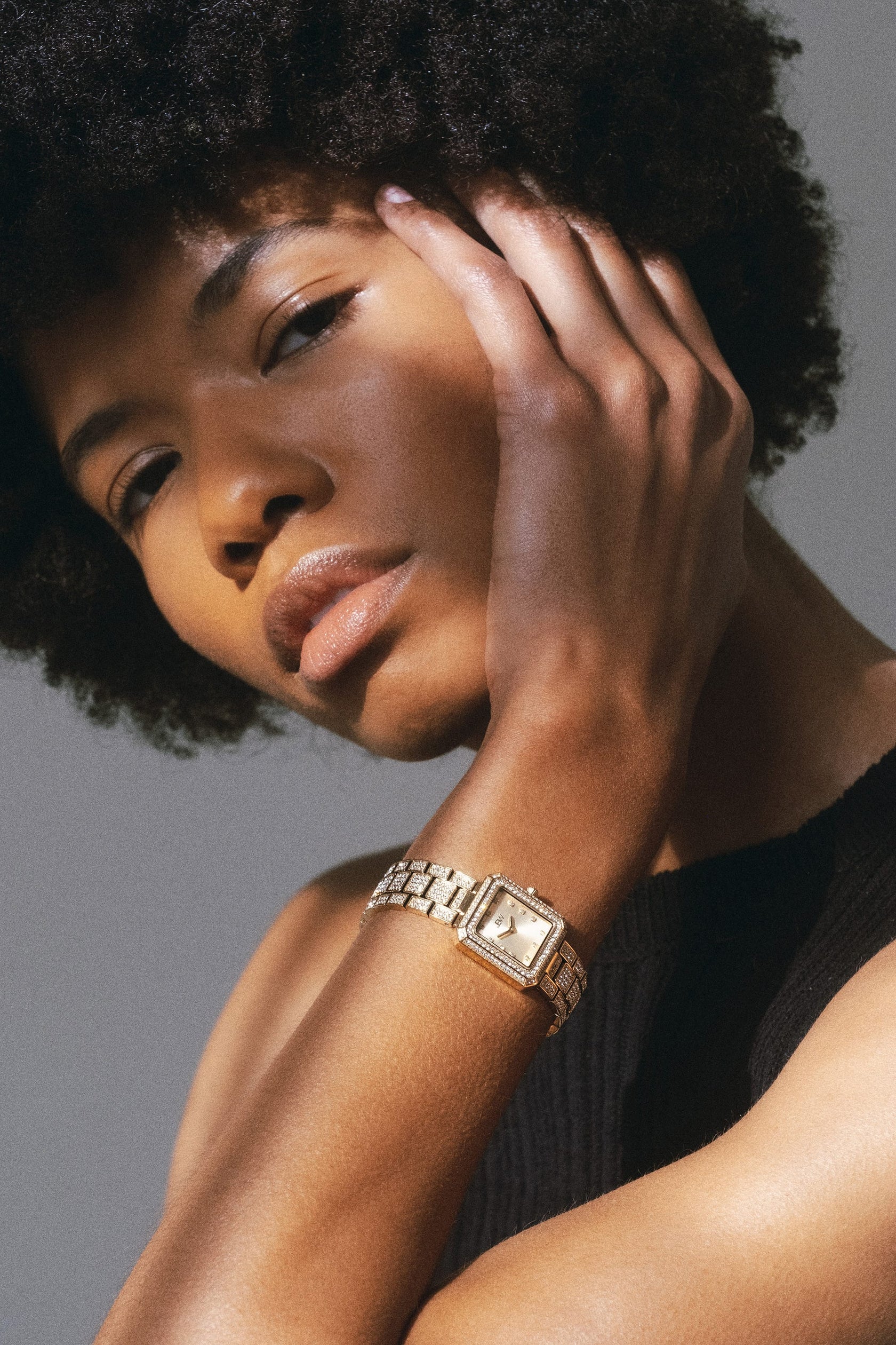 A person with short curly hair poses with one hand on their face, wearing a sleeveless black top and the JBW Watches Arc Single (Sample Sale) | J6390A-GA, its crystal accents catching the soft light that highlights their smooth skin and thoughtful expression.