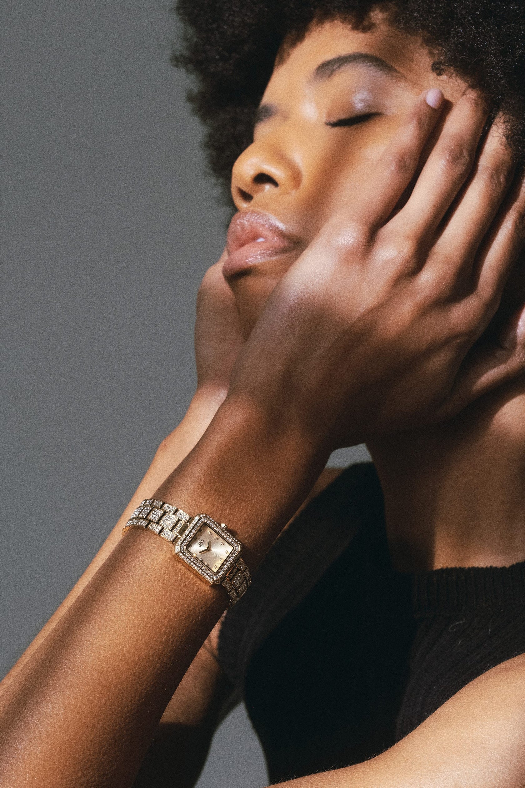 With a calm, contemplative mood, a person in a sleeveless black top gently touches their face, showcasing the JBW Watches Arc Single (Sample Sale) | J6390A-GA unisex crystal-embellished square-face watch on their left wrist.