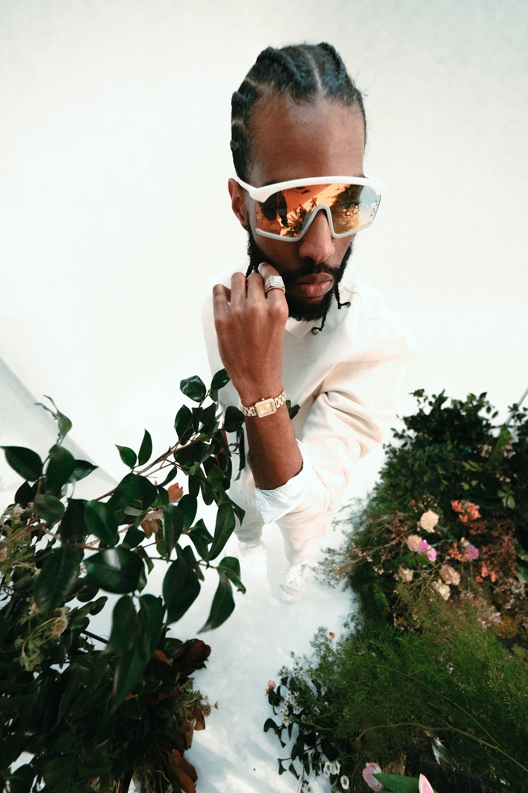 A man with braided hair, reflective sunglasses, a white shirt, crystal-embellished jewelry, and a JBW Watches Arc Single (Sample Sale) watch poses confidently among lush plants and flowers against a neutral background.